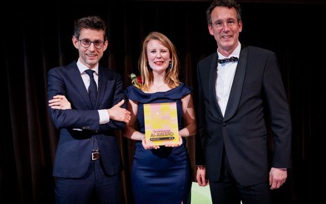 Een vrouw met donkerblauwe galajurk houdt een plaquette vast waarop staat: De Nederlandse AI-Award. Links van haar staat een man met een donkerblauw pak en een stropdas, rechts van haar staat een man met een antraciet pak en een strikje. 