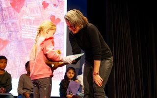 Een vrouw geeft een boek aan een meisje, op de achtergrond meerdere kinderen die boeken vasthouden.