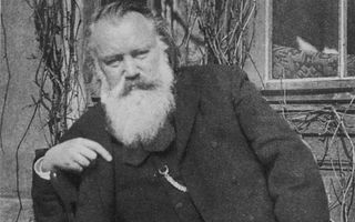 Een oudere man met een grote witte baard en een donker pak aan staat voor takken waaraan geen blaadjes zitten.