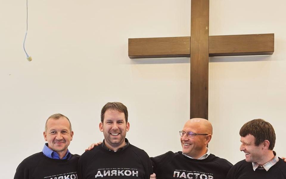 Vier lachende mannen in zwarte truien met witte letters erop staan met de armen om elkaar heen geslagen.