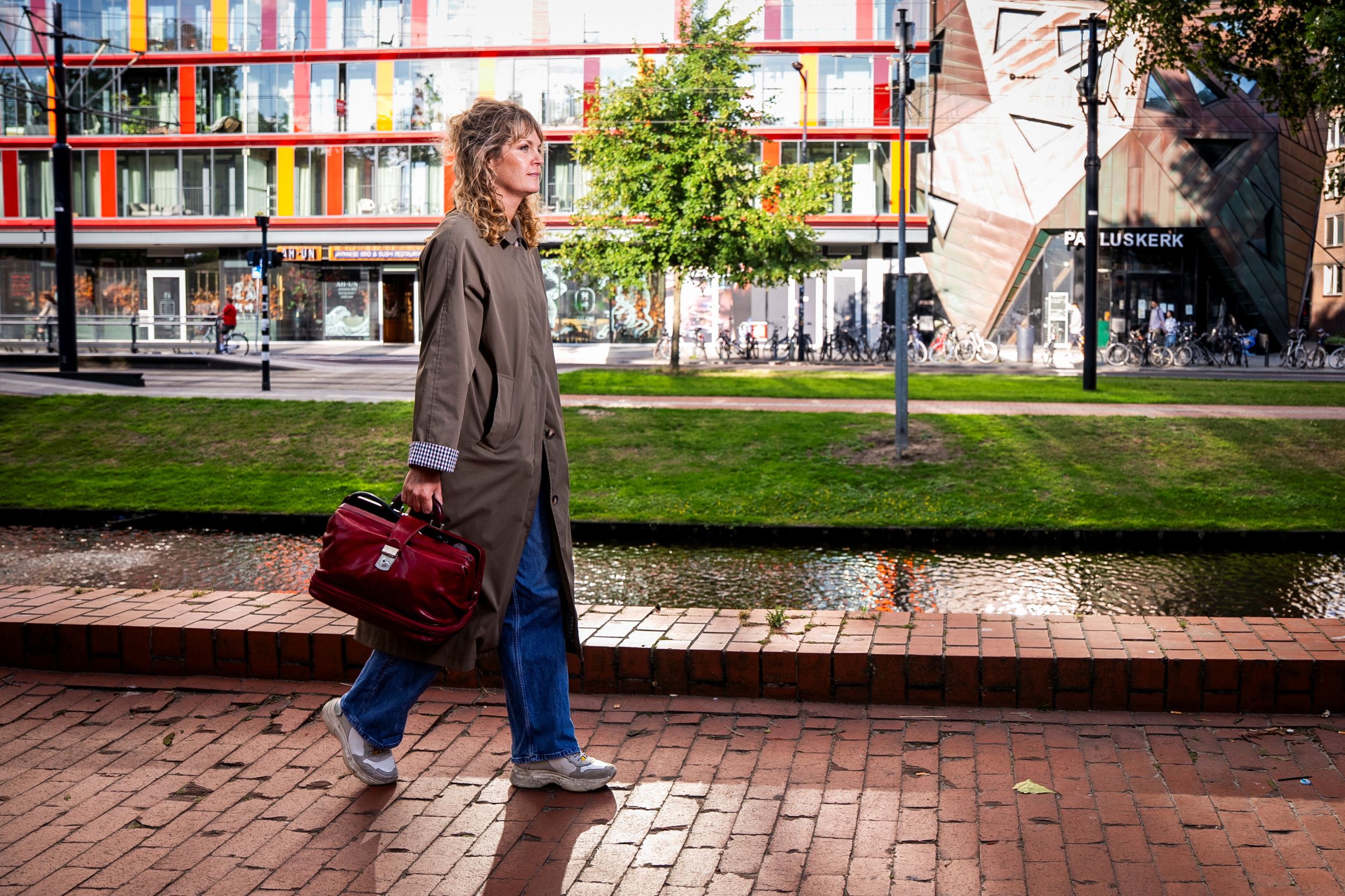 Vrouw met bruine jas en rode dokterstas loopt over een glimmend natte stoep van rode klinkers. Aan de overkant een bruin, hoekig gebouw met ”Pauluskerk” erop, naast een gebouw met vanaf de eerste verdieping rode en oranje vakken naast ramen. 