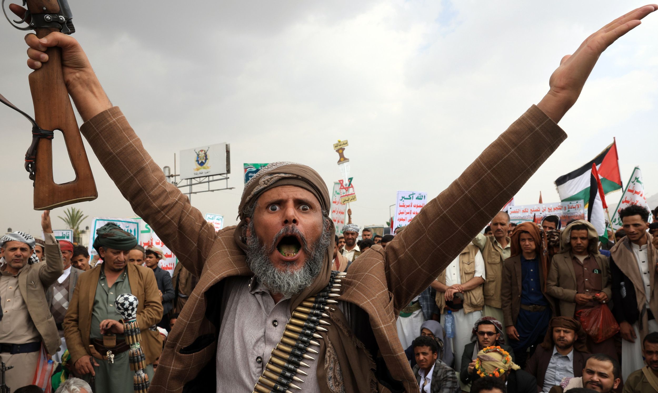 Man met baard in bruin jasje en omhangen met kogels, steekt een geweer in de lucht.