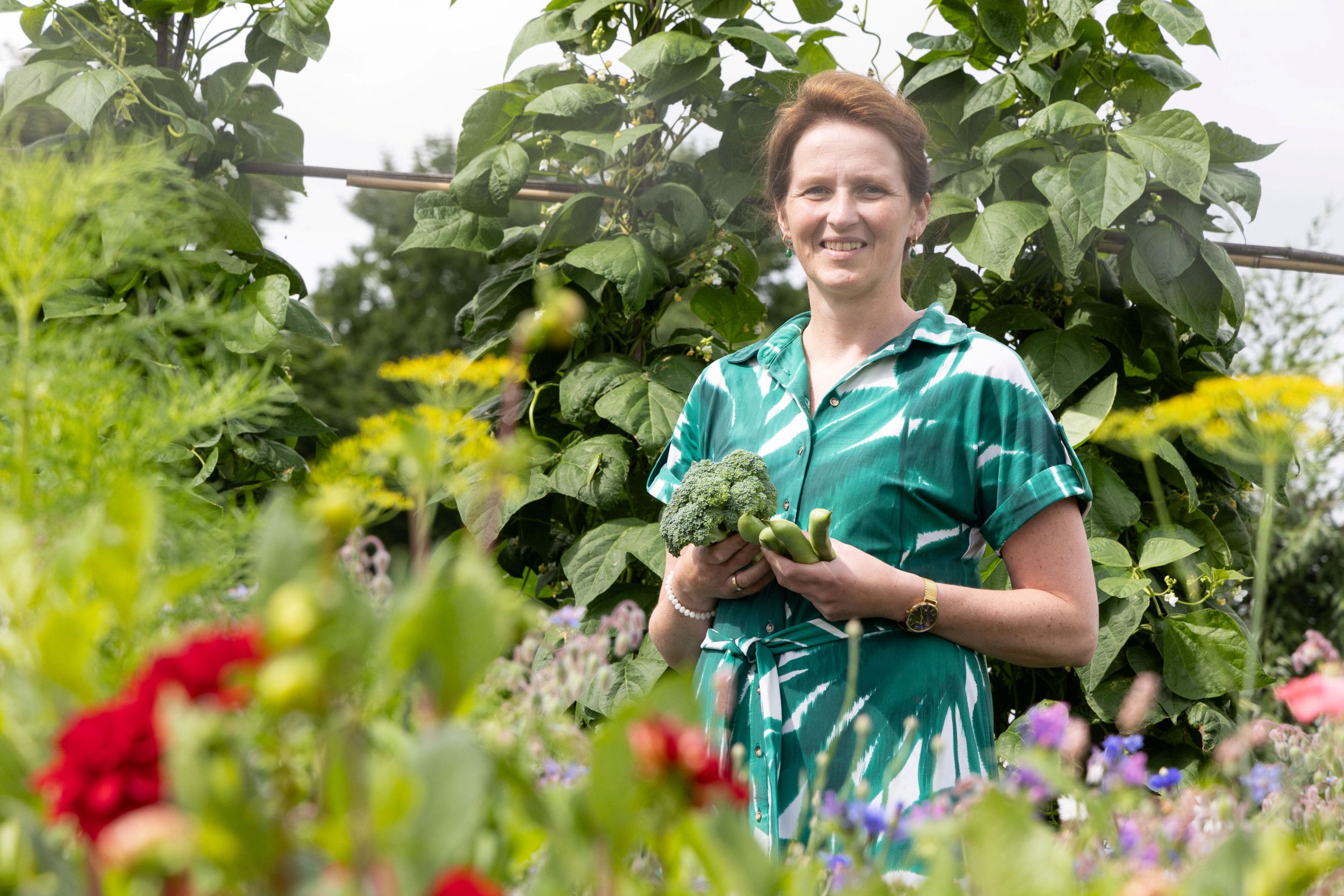 Een vrouw in een groen-witte jurk staat in een groentetuin. In haar hand wat boontjes en broccoli. Op de achtergrond allerlei groenten, op de voorgrond wat bloeiende bloemen, geel, rood en roze. 