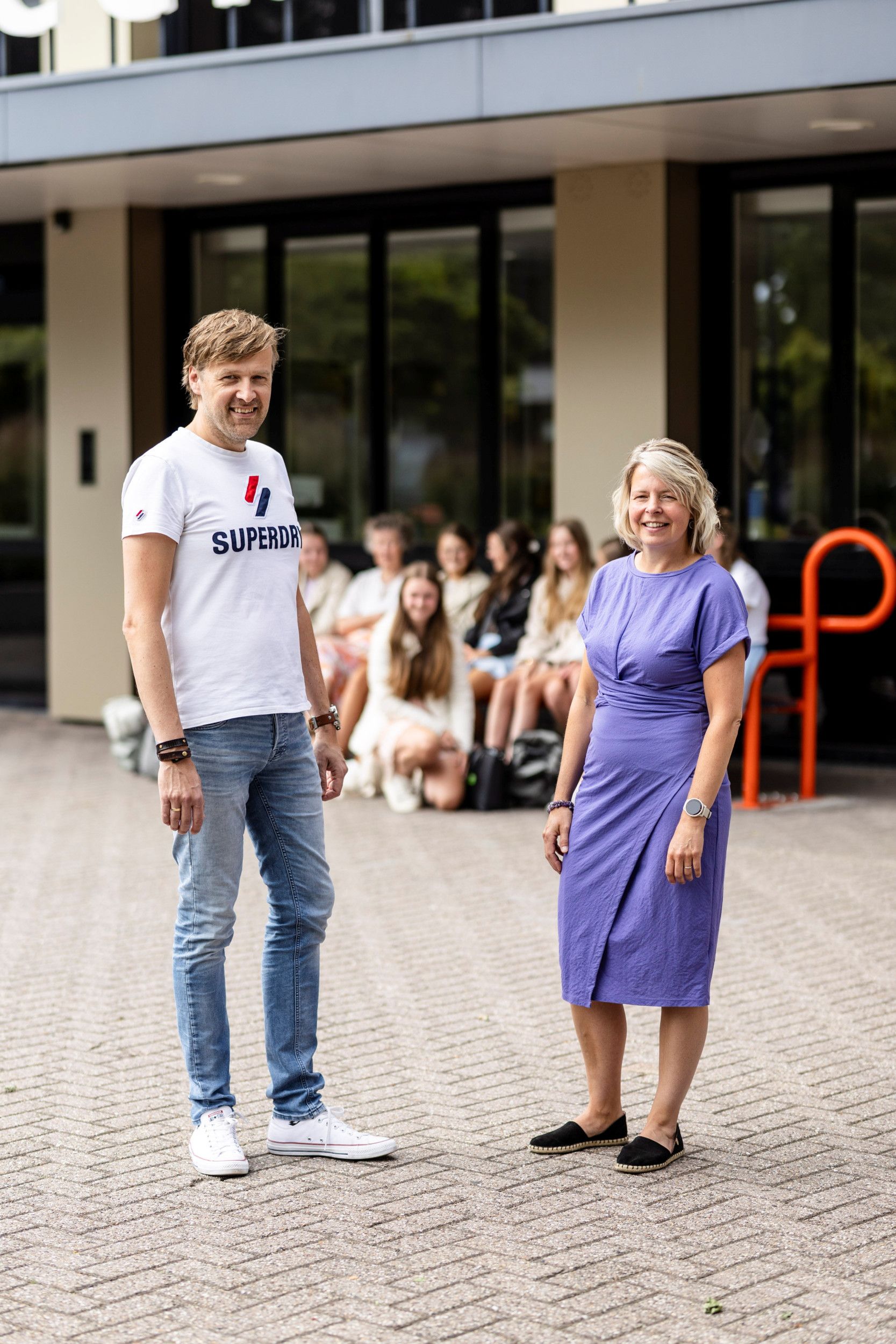 Een man in een wit t-shirt en een spijkerbroek staat op het schoolplein. Naast hem een vrouw in een paarse jurk en zwarte schoenen. 