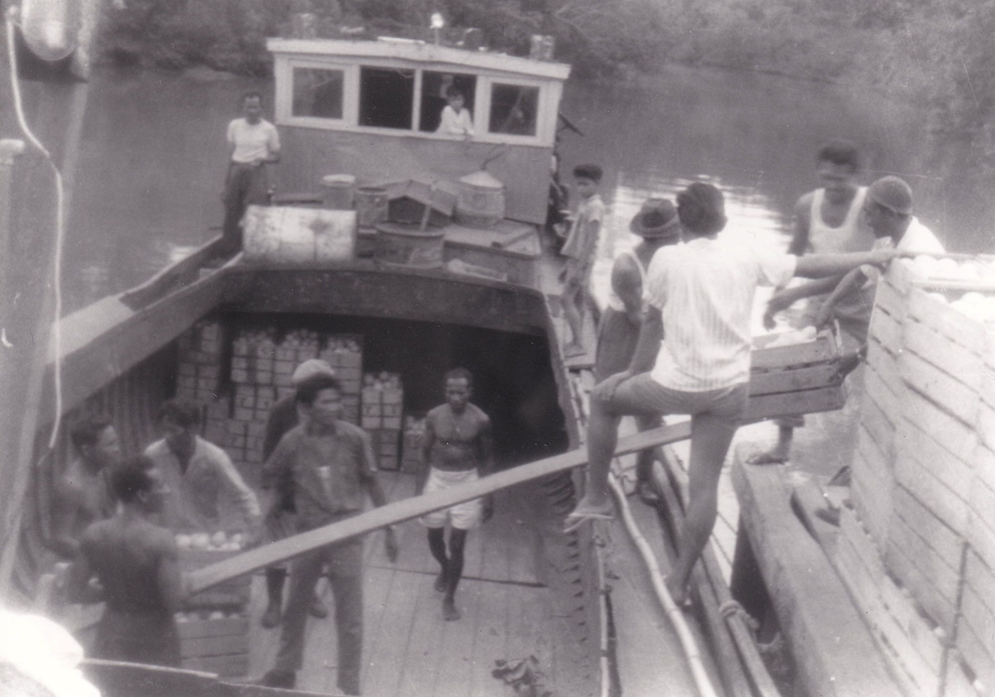 Donkere mannen laden kisten vol citrusvruchten vanaf een steiger in het ruim van een klein vrachtschip. Ze laten de kisten over een smalle plank glijden.