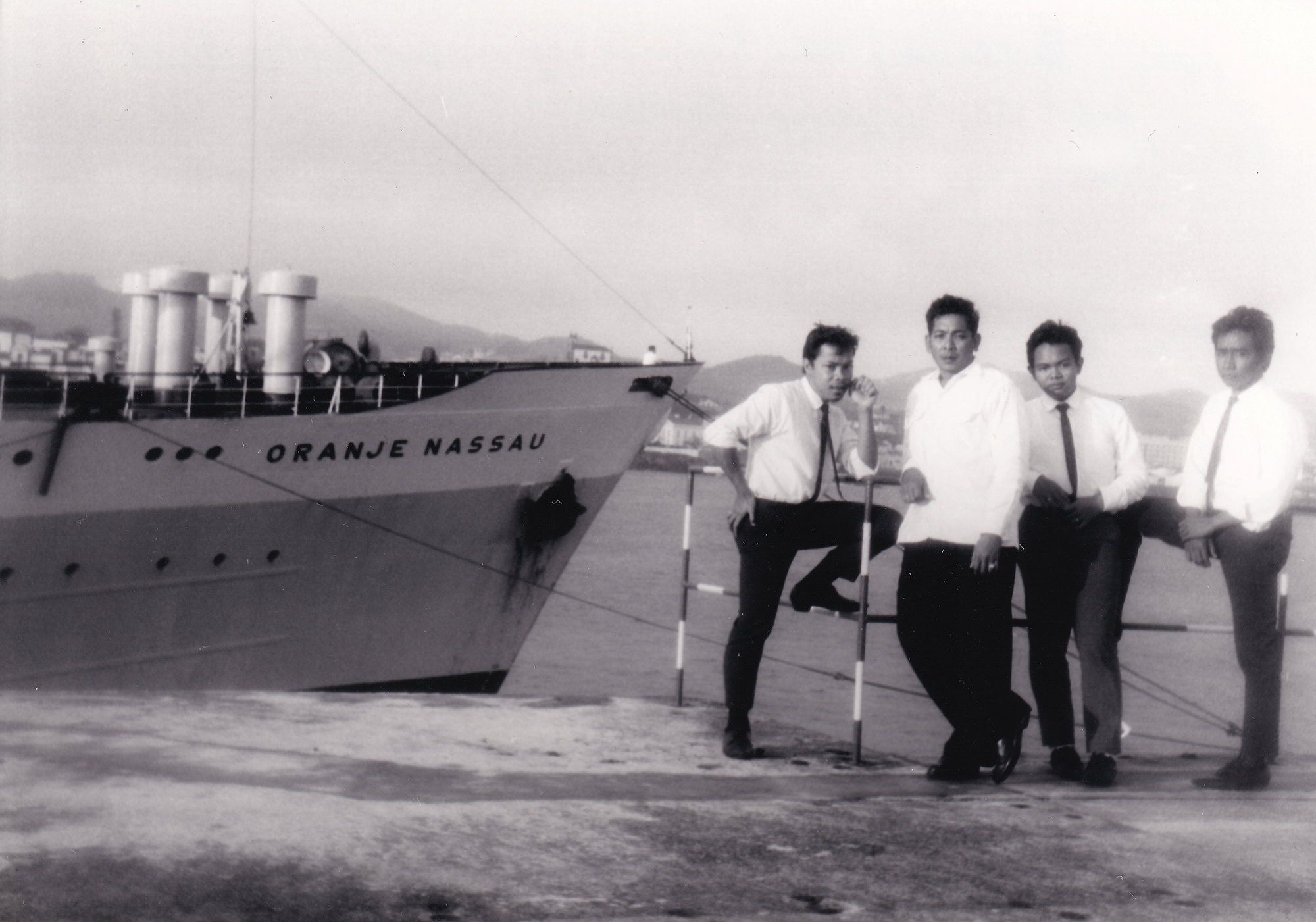 Vier donkere mannen met zwarte broek en wit overhemd staan aan de kade. Links is de boeg van een schip te zien met op de steven de naam ”Oranje Nassau”. Op de achtergrond zijn bergen te zien.