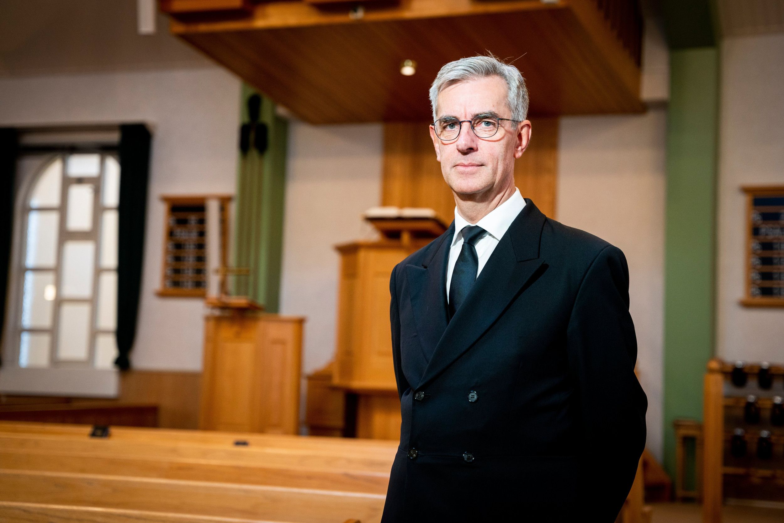 Predikant van middelbare leeftijd met bril en grijs haar in zwart pak met zwarte stropdas, gefotografeerd in kerkgebouw met houten banken en preekstoel.