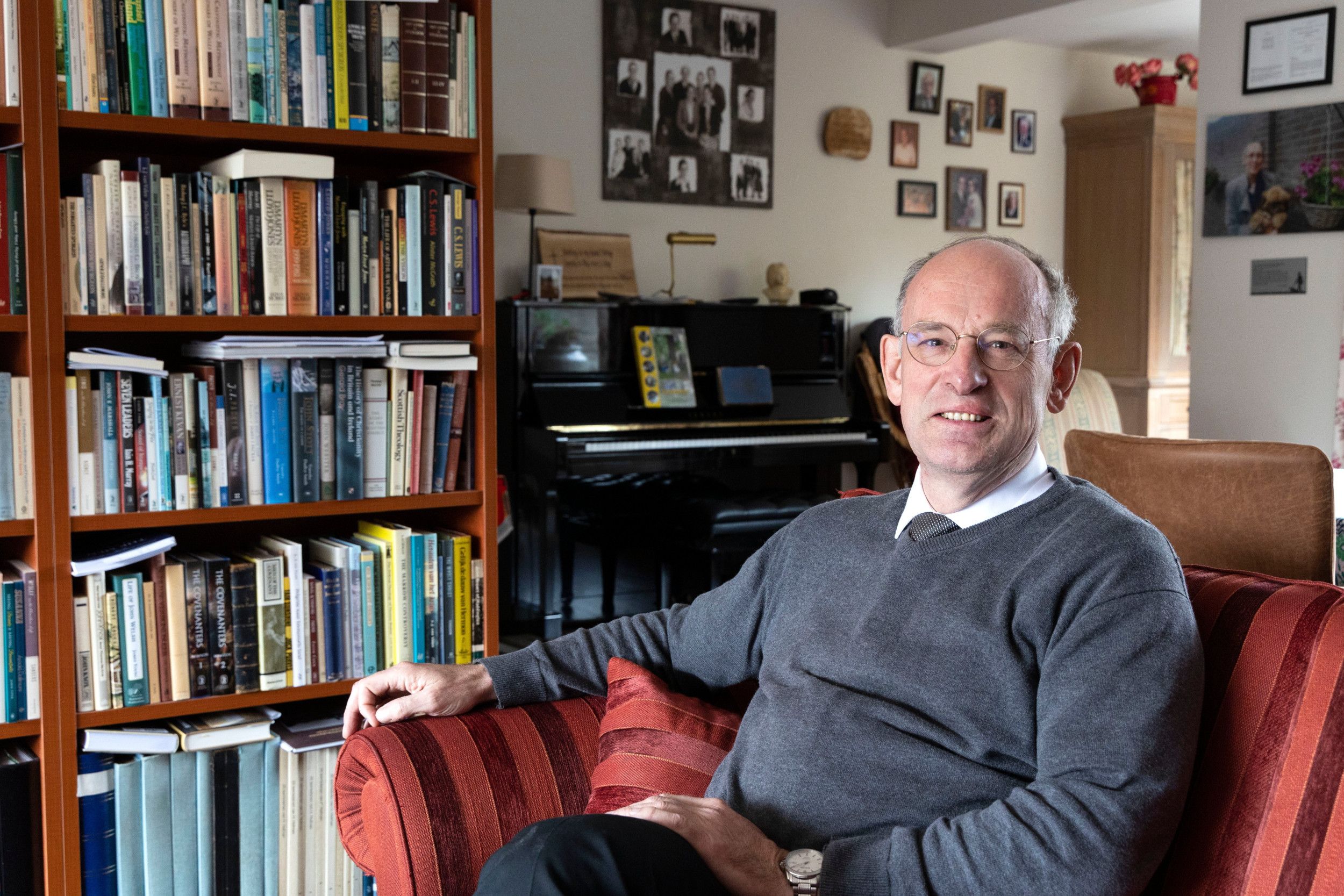 Predikant met bril in donkergrijze trui met stropdas zit in rode stoel naast boekenkast en piano. De muur hangt vol foto’s.