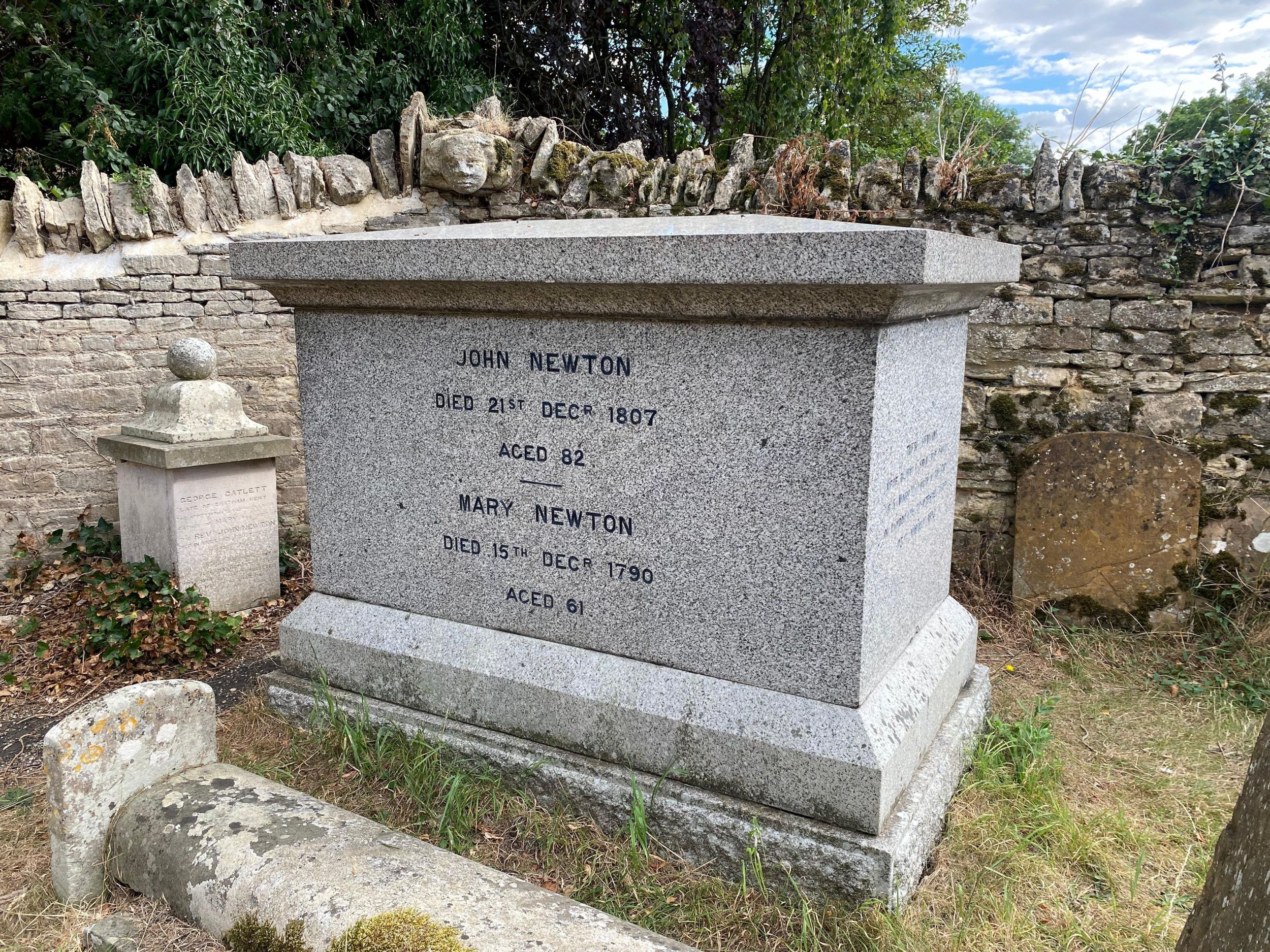 Grijze graftombe met zwarte letters. Op de achtergrond andere grafzerken en vervallen stenen muur.