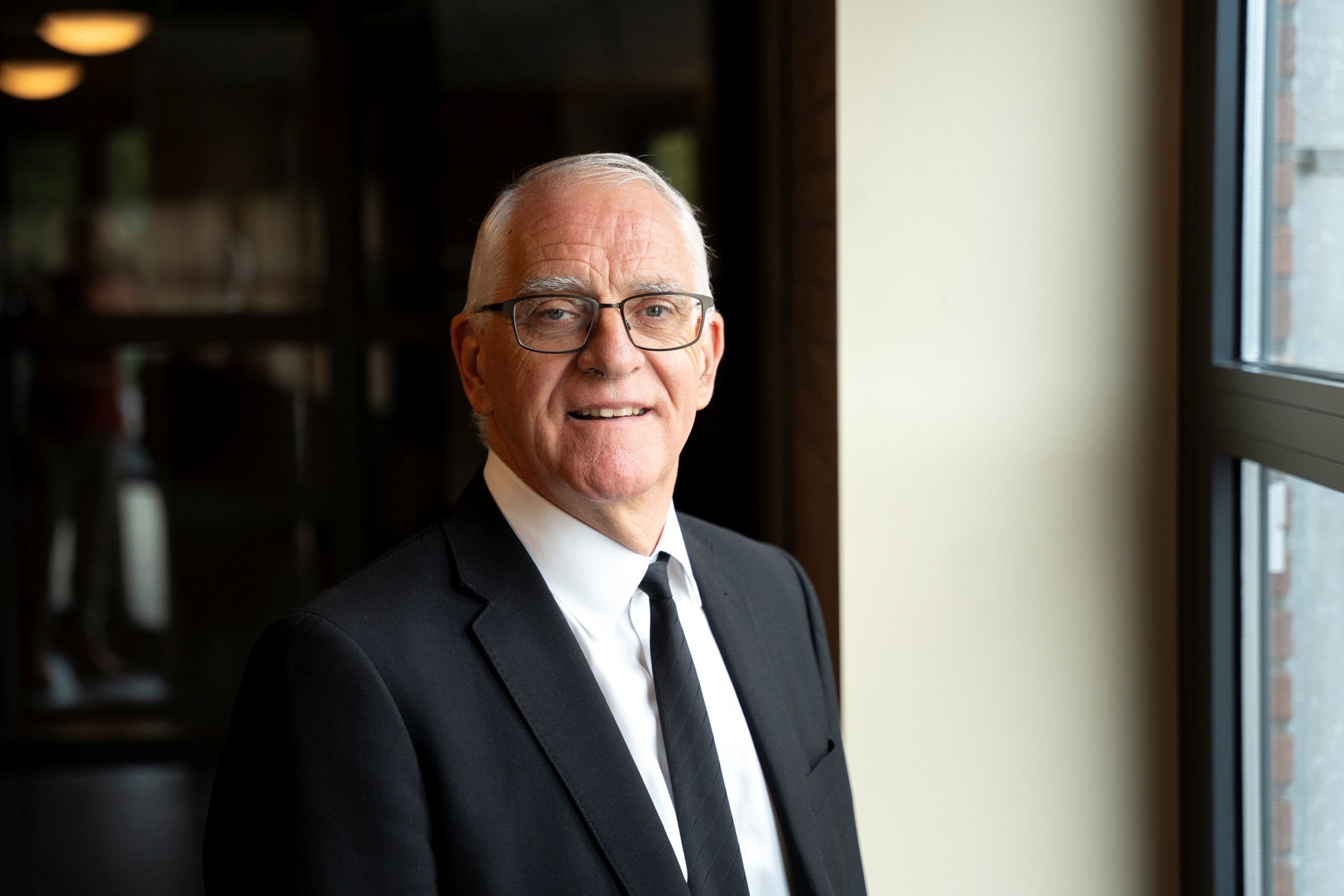 Dominee Pocock, een man met grijs haar, bril op en gekleed in een zwart pak.