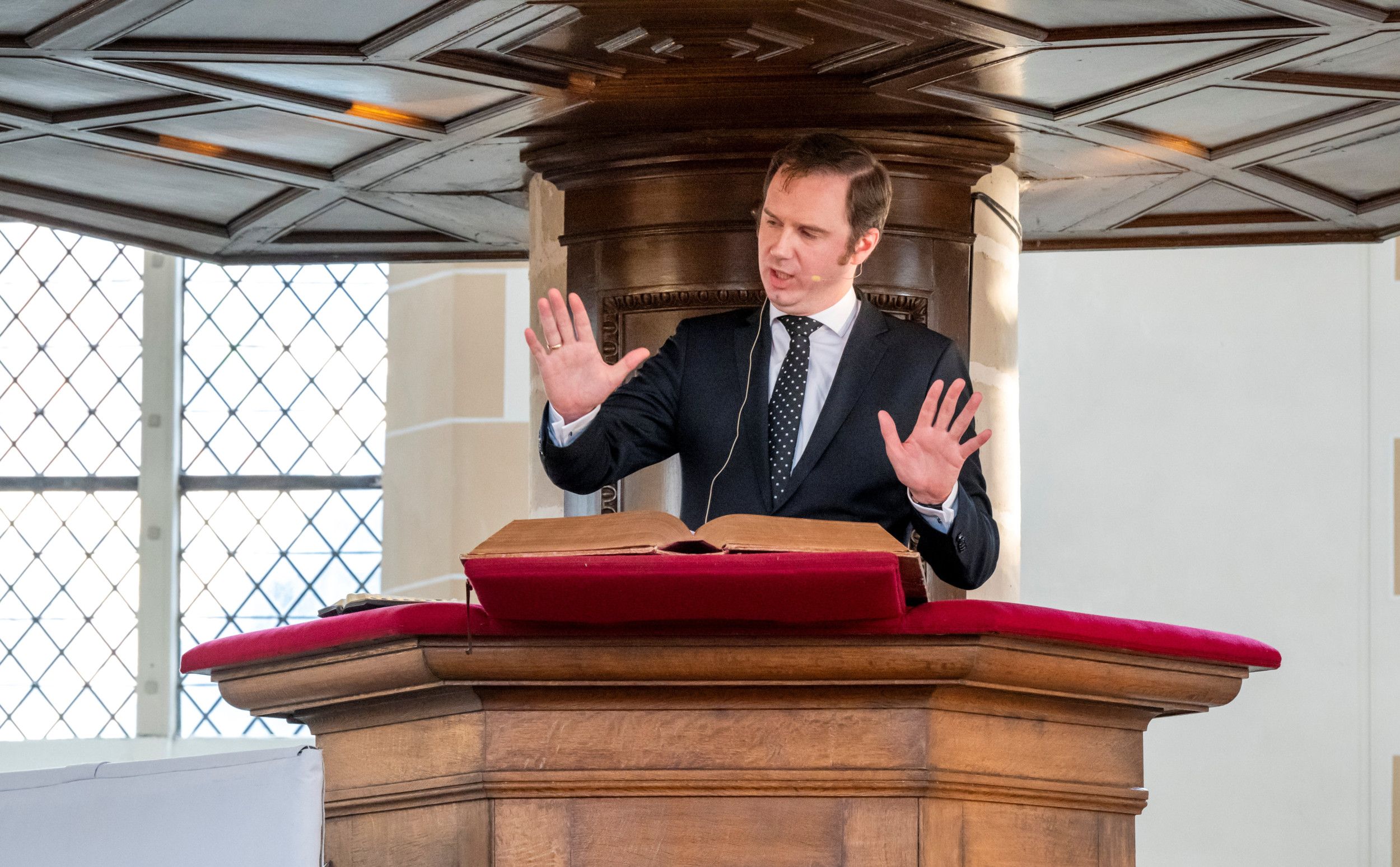 Dominee Fokkema, in zwart kostuum, staat op een kansel, met opgeheven handen.