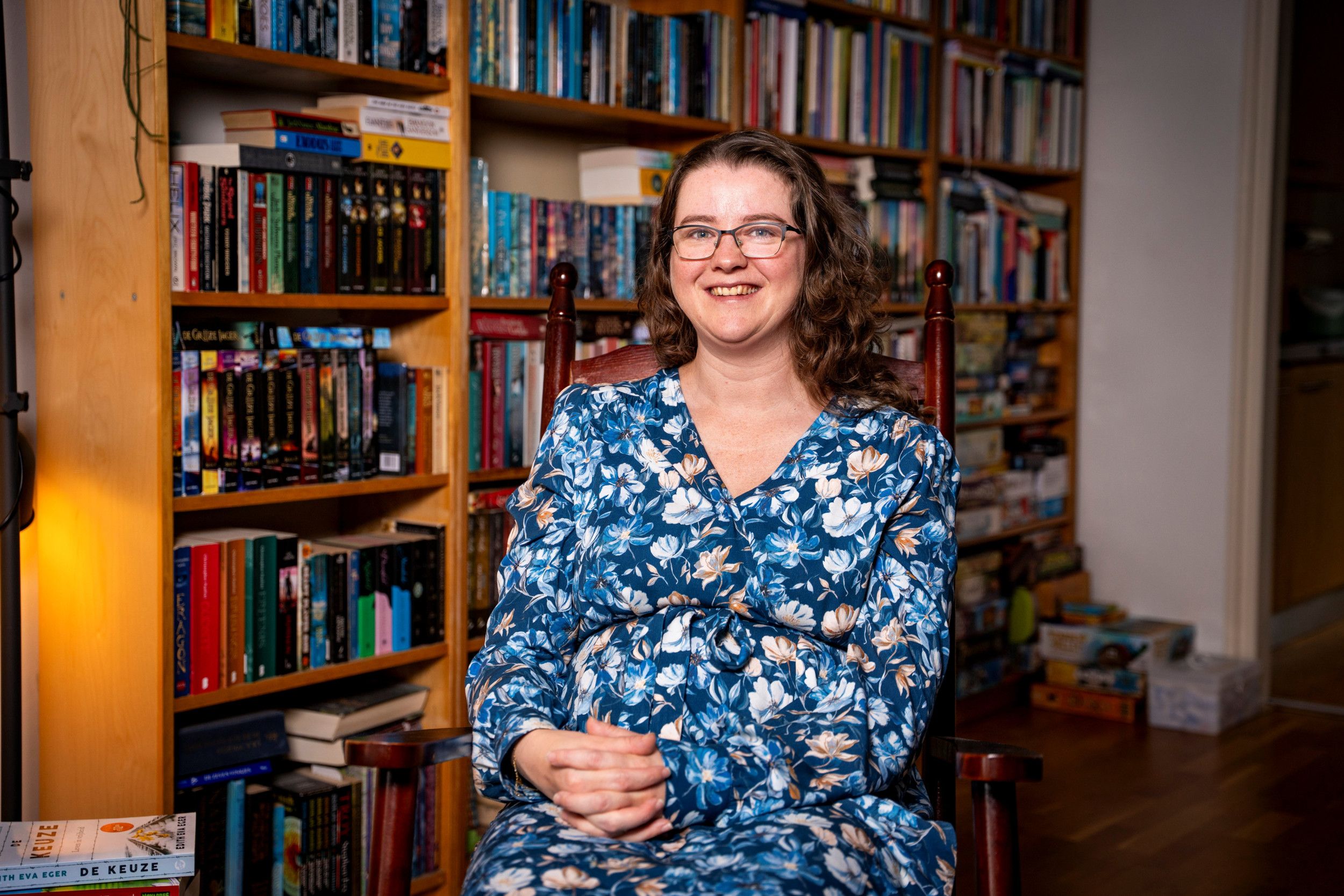 Vrouw in blauwe bloemenjurk zit voor een grote boekenkast.