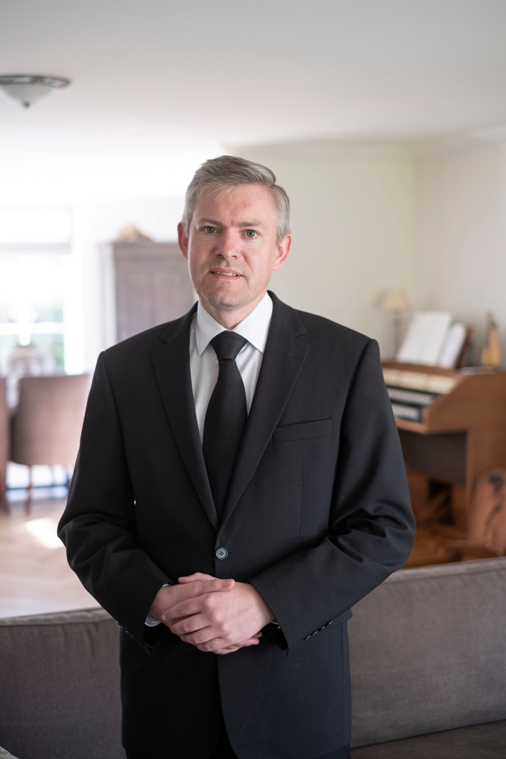 Predikant in zwart pak in zijn woonkamer, orgel op de achtergrond.