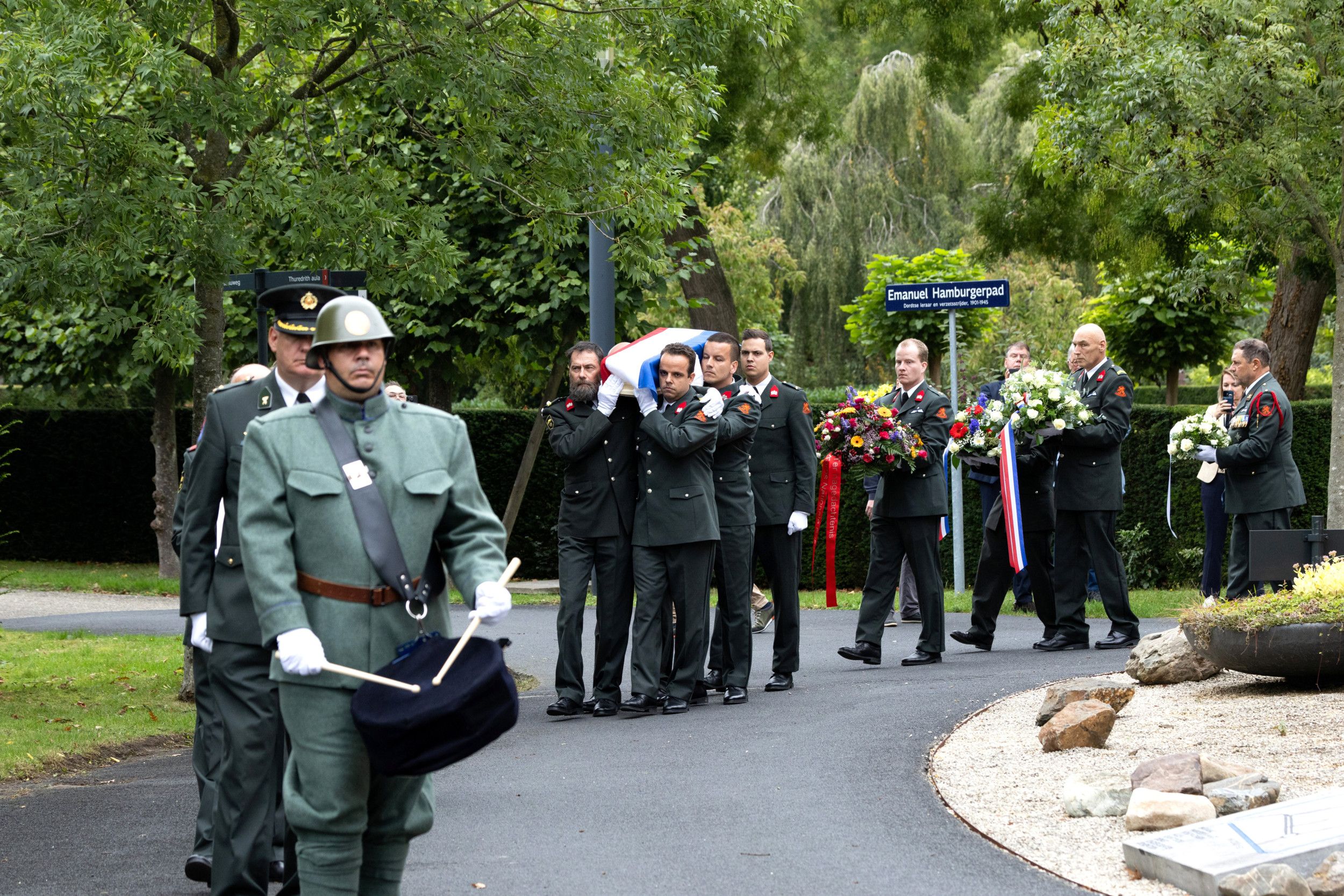 Militairen met trom, met een kistje waarop een vlag op de schouders, en met bloemstukken.