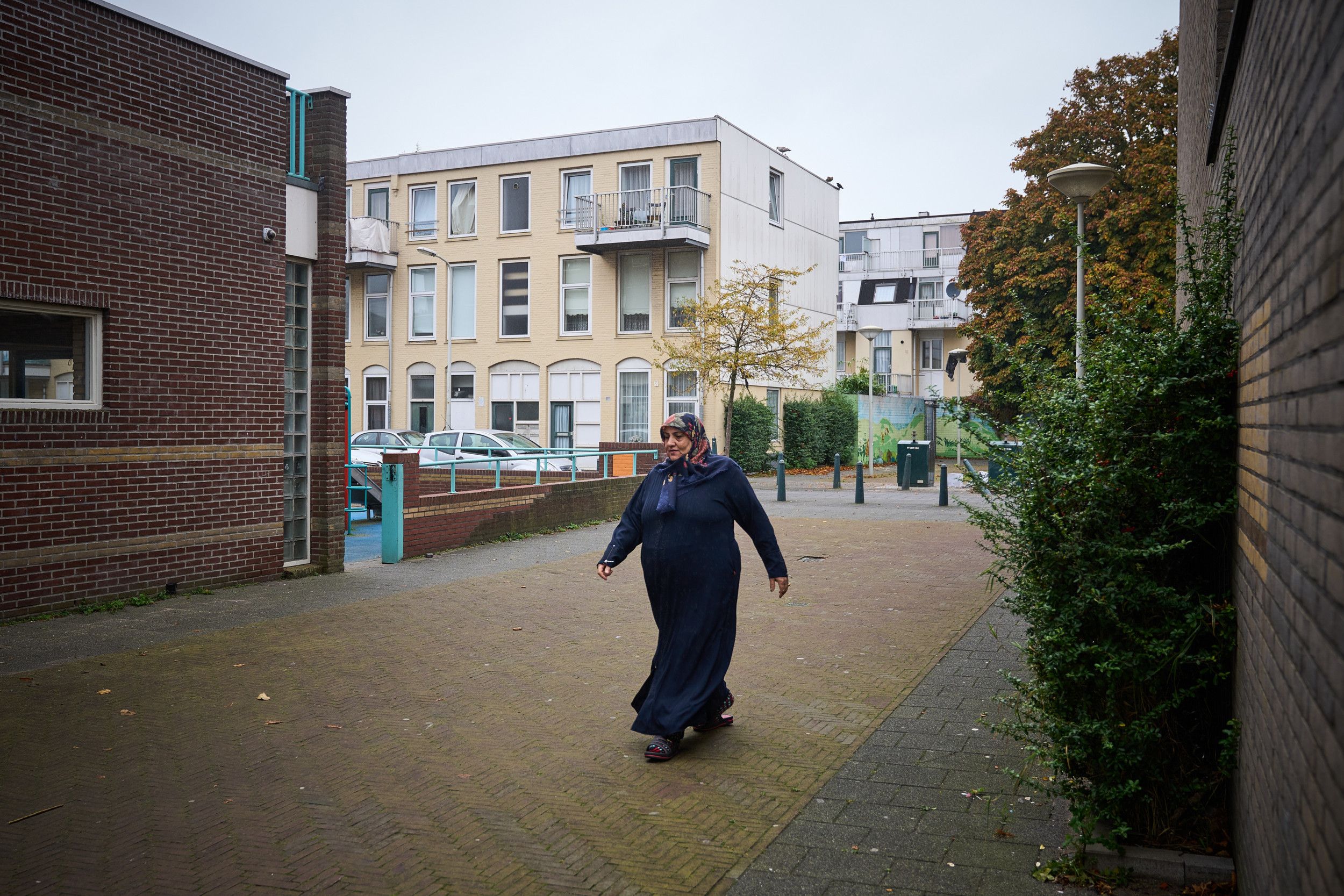 Een vrouw met een donkerblauwe jurk en hoofddoek loopt door een klinkerstraatje met een flat met huurwoningen op de achtergrond. 