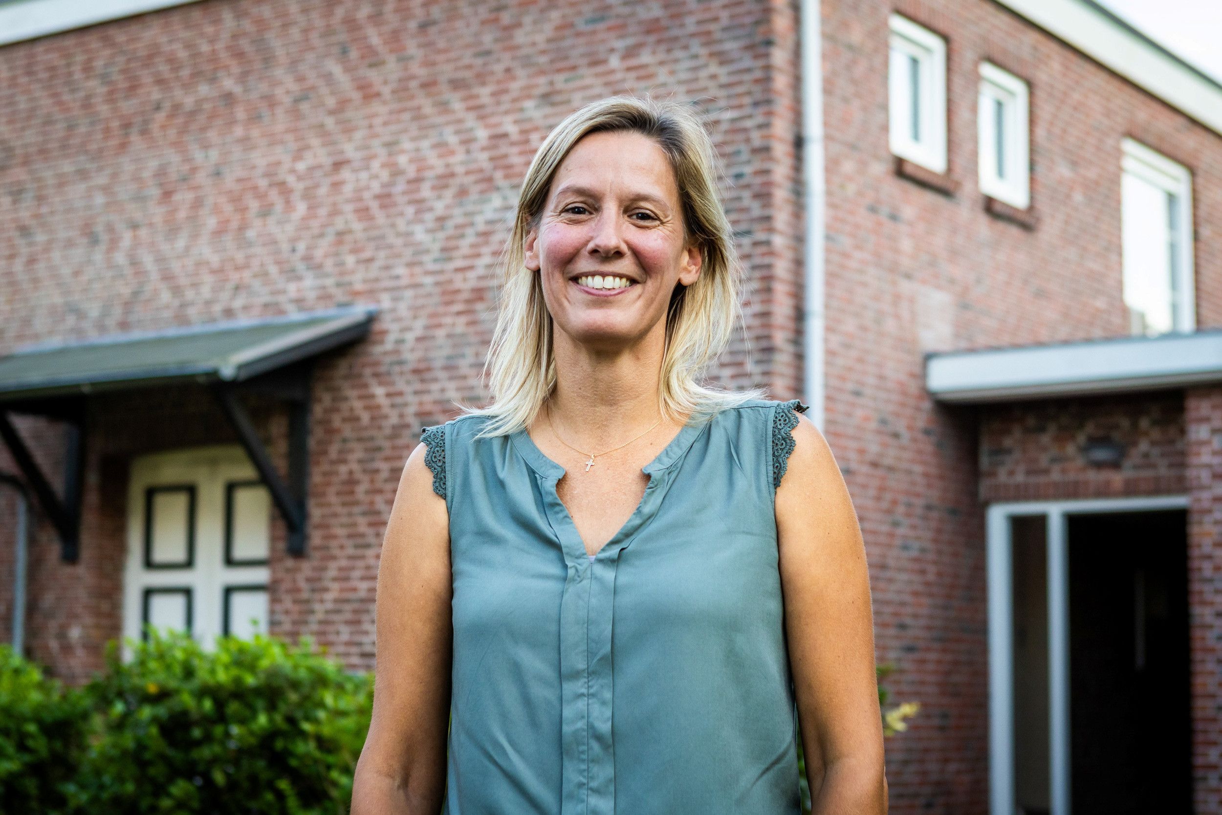 Vrouw van middelbare leeftijd met blond haar en een blauwe blouse kijkt lachend in de camera. 