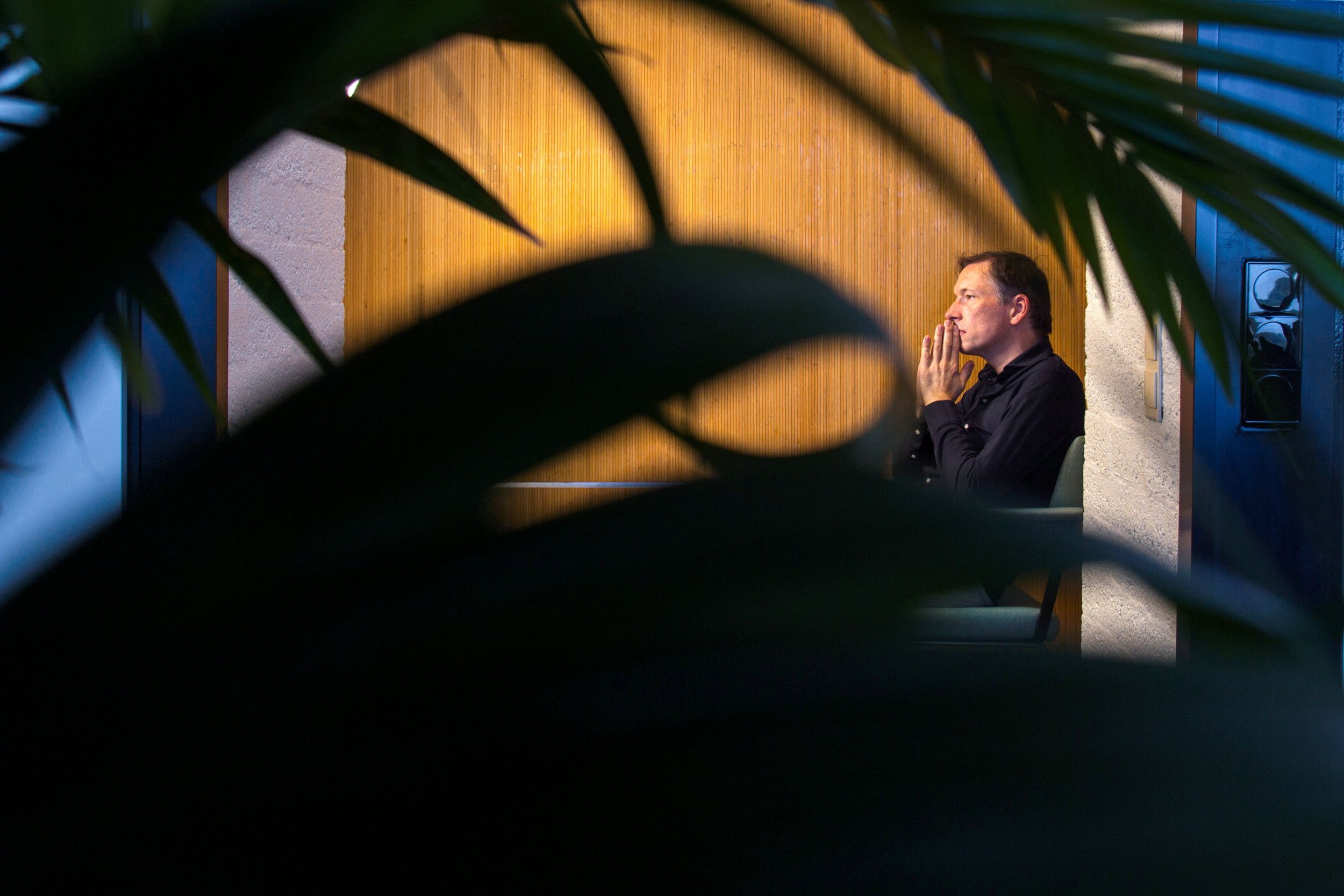 Man van middelbare leeftijd met donkerblauw overhemd zit op een stoel; hij heeft zijn handen tegen elkaar gedrukt tegen zijn kin aan. De man staart in de verte. Je ziet hem tussen de bladeren van een plant door.