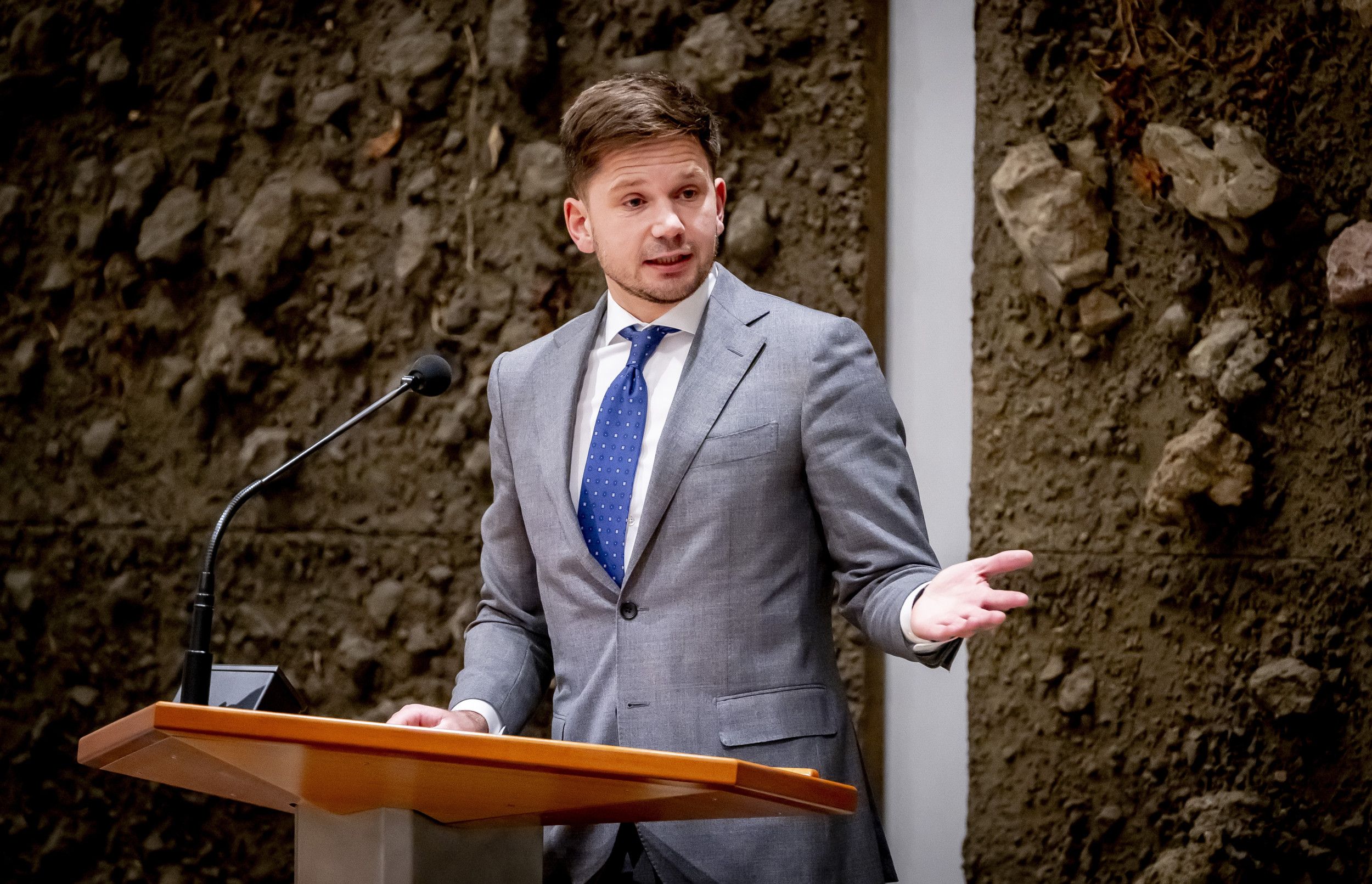 Man in pak achter het spreekgestoelte voor Kamerleden in de plenaire zaal van de Tweede Kamer.