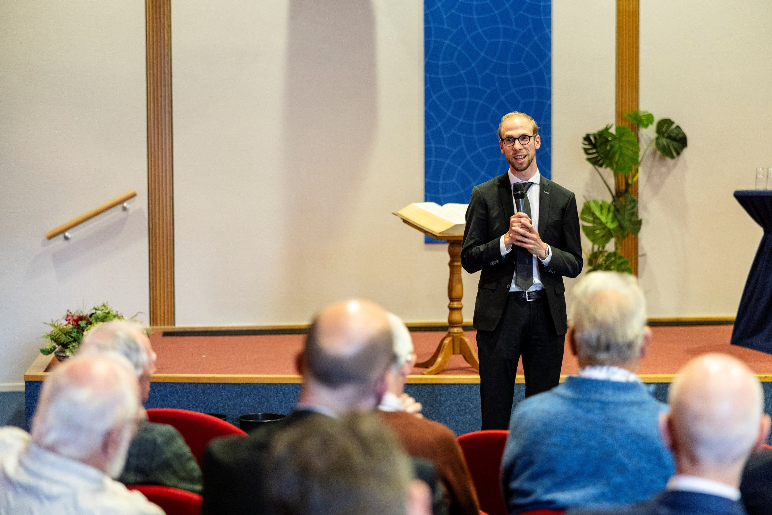 Man met bril en baardje, in donker pak, staat voor in een zaal, met microfoon in de hand.