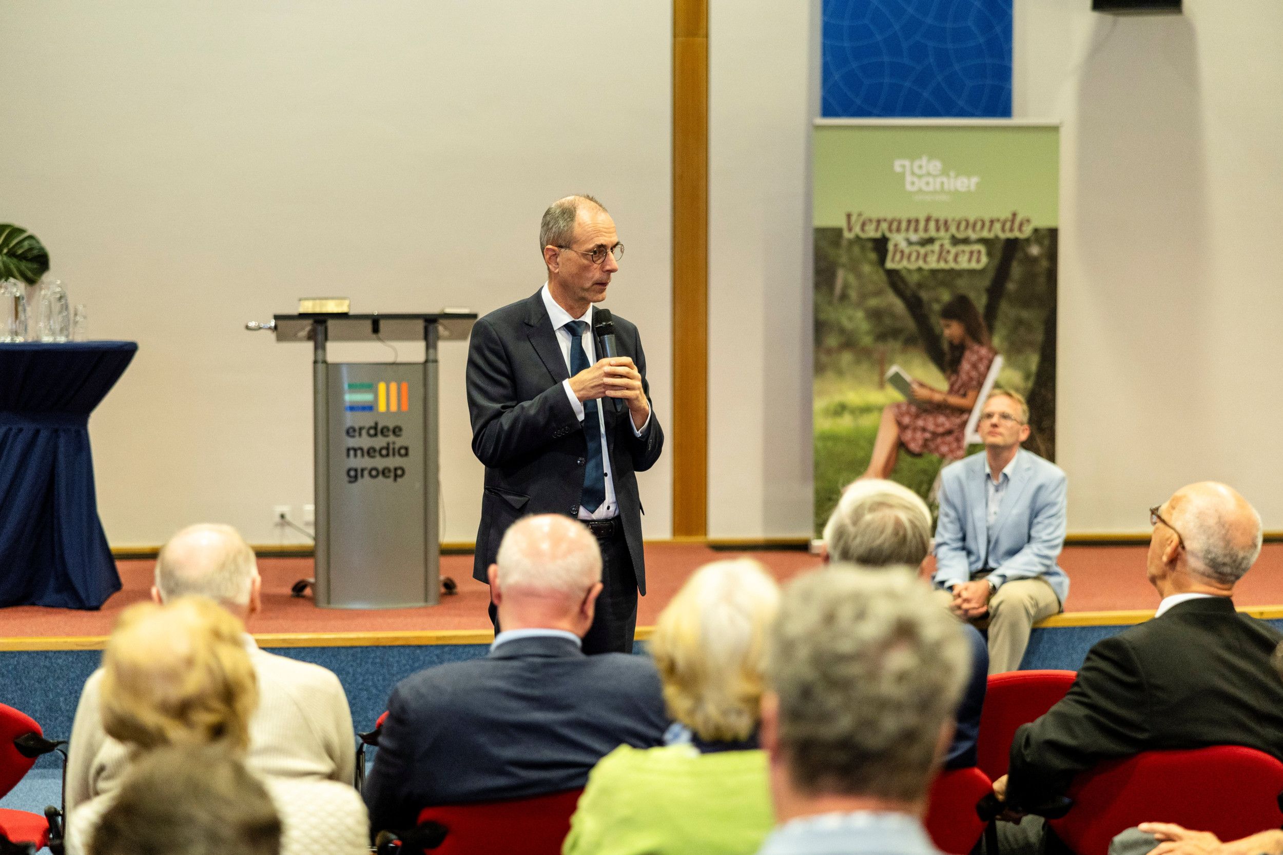 Man met bril, in donker pak, staat voor in een zaal, met microfoon in de hand.