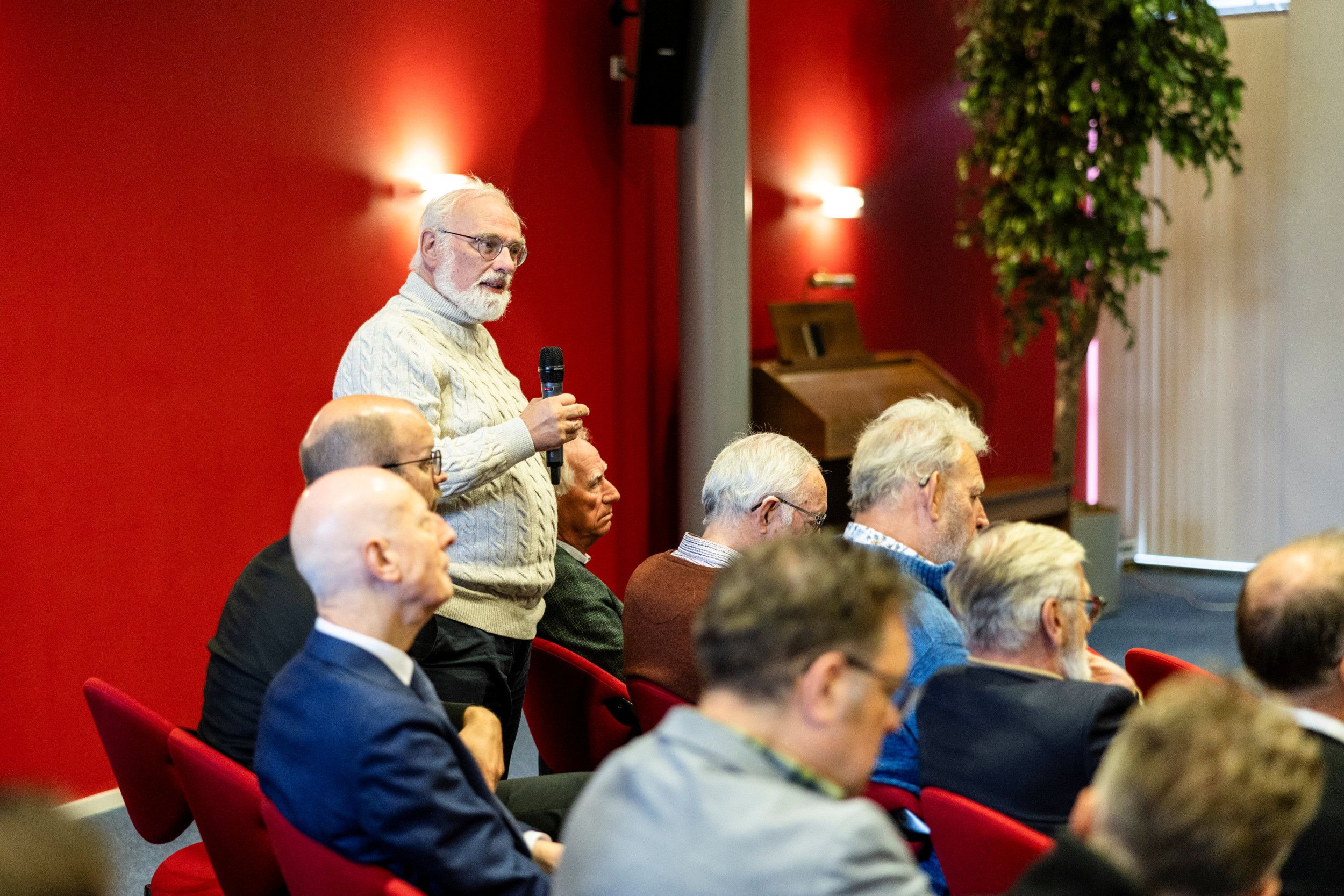 Man met witte baard, in witte wollen trui, met microfoon in de hand.