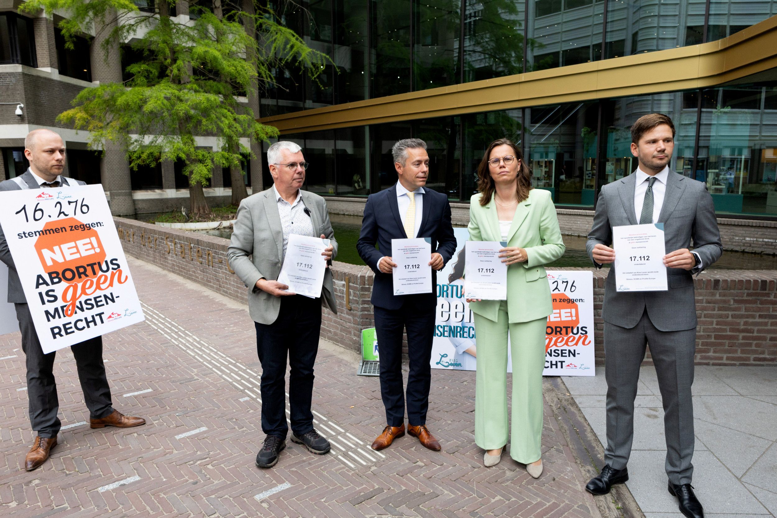 Vijf mensen staan buiten voor het gebouw van de Tweede Kamer. Een van hen heeft een bord met een tekst voor zijn buik, de andere vier hebben een document in hun handen.