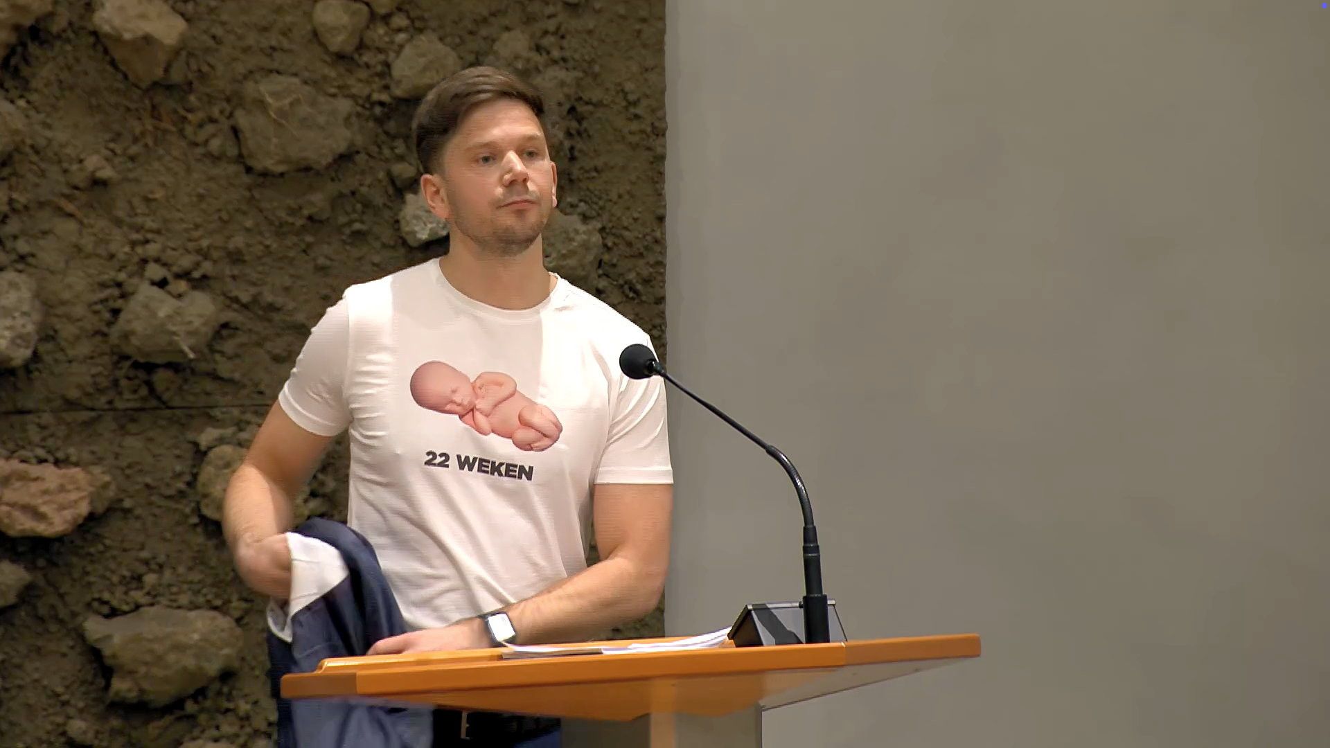 Man achter spreekgestoelte in de Tweede Kamer doet zijn jasje uit, waaronder hij een T-shirt met daarop de afbeelding van een foetus draagt.