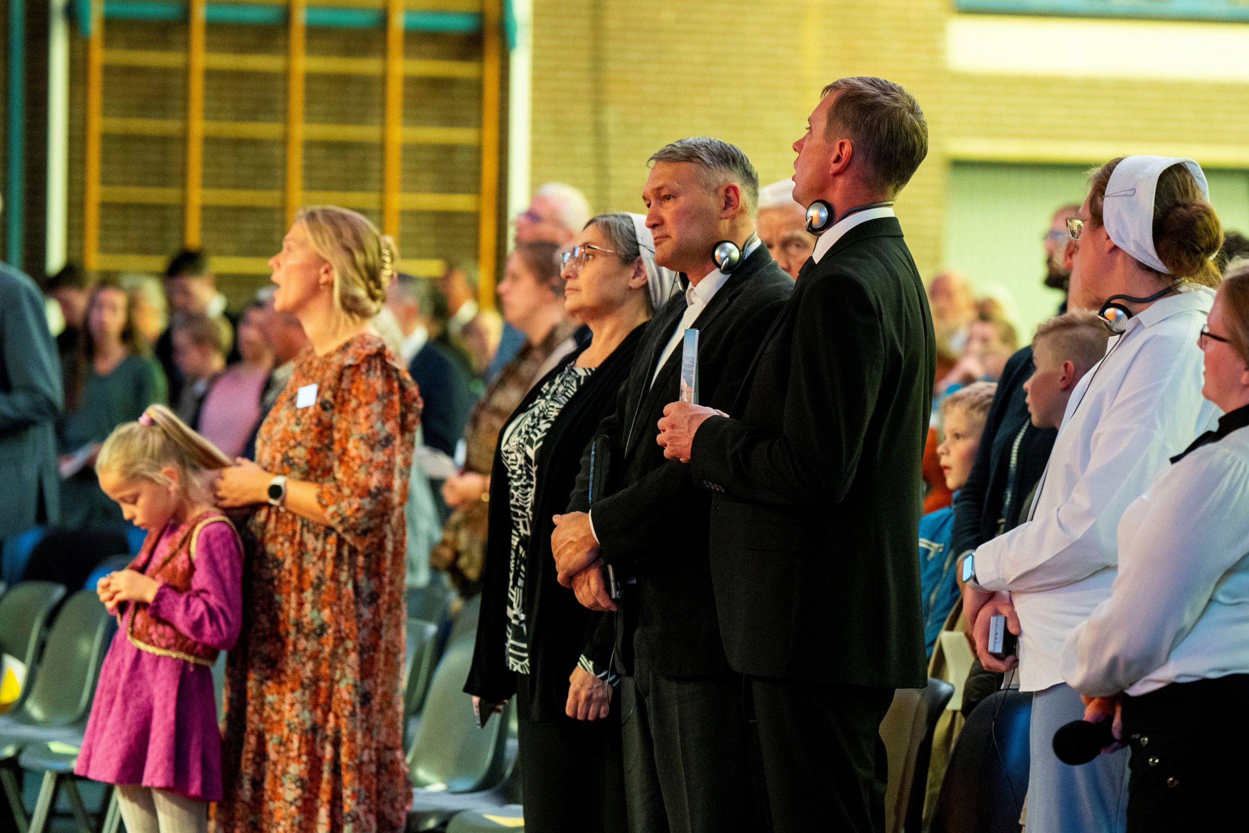Beeld van zingende mensen in een grote gymzaal waarin stoelen staan opgesteld. In het midden een in donkere kleding gekleed echtpaar, de Poegatsjevs. 