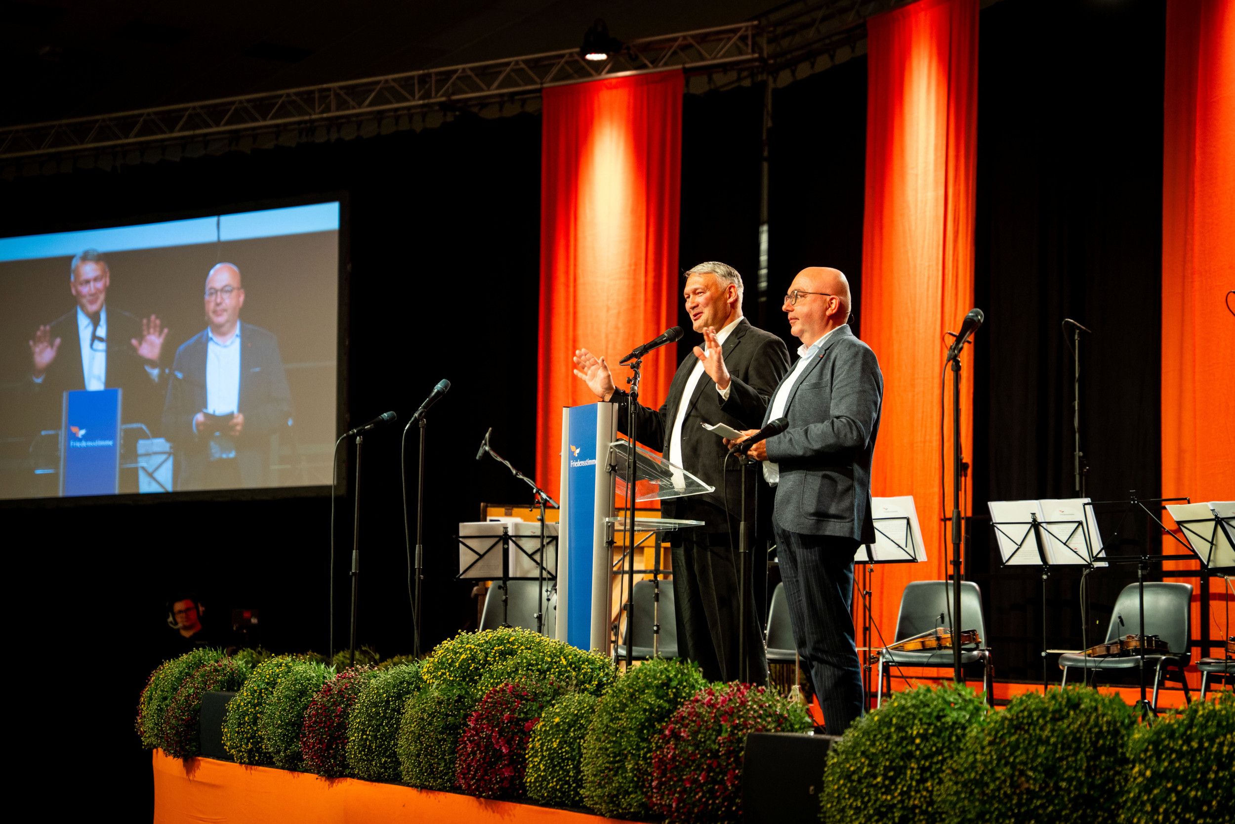 Podium waarop twee mannen in pak staan, eromheen muziekstandaarden en microfoons, op de voorgrond bolchrysanten. 