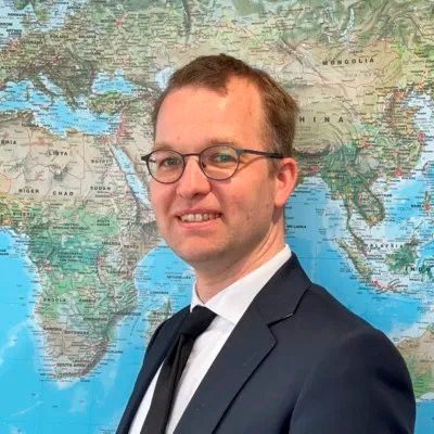 Man met bril in zwart pak met stropdas met wereldkaart op achtergrond.