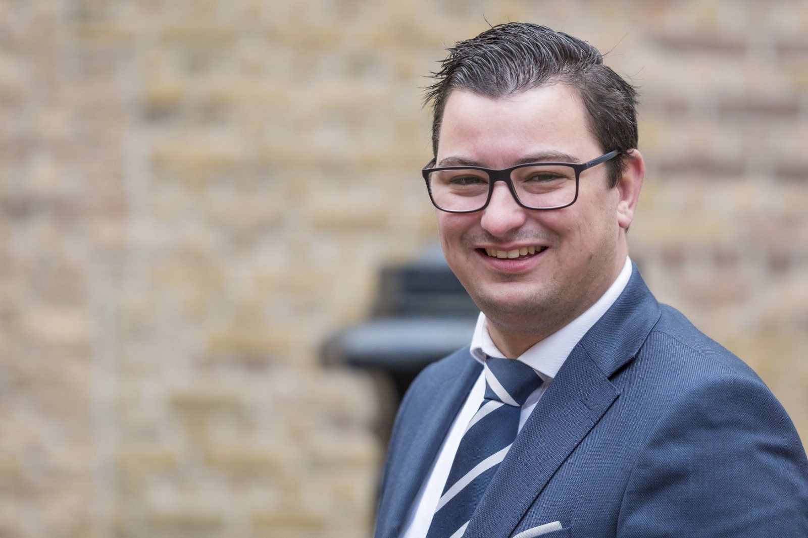 Lachende man met donker haar en een bril. Hij draagt een pak met gestreepte stropdas.