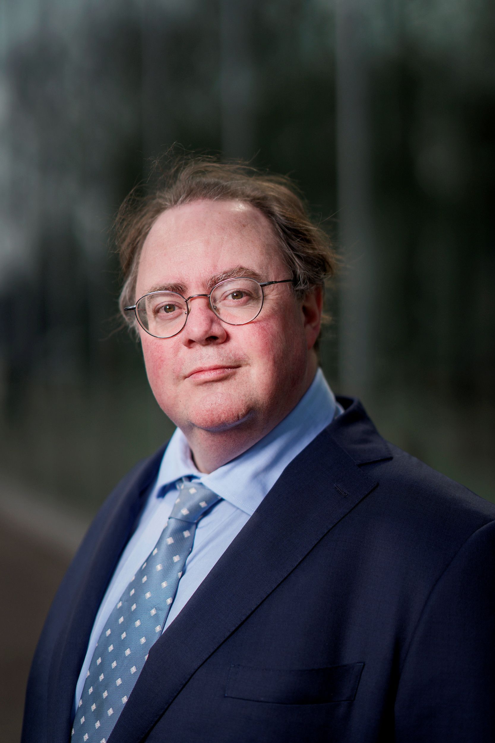 Een man met bril, stropdas en donkerblauw jasje kijkt in de camera voor een portretfoto.