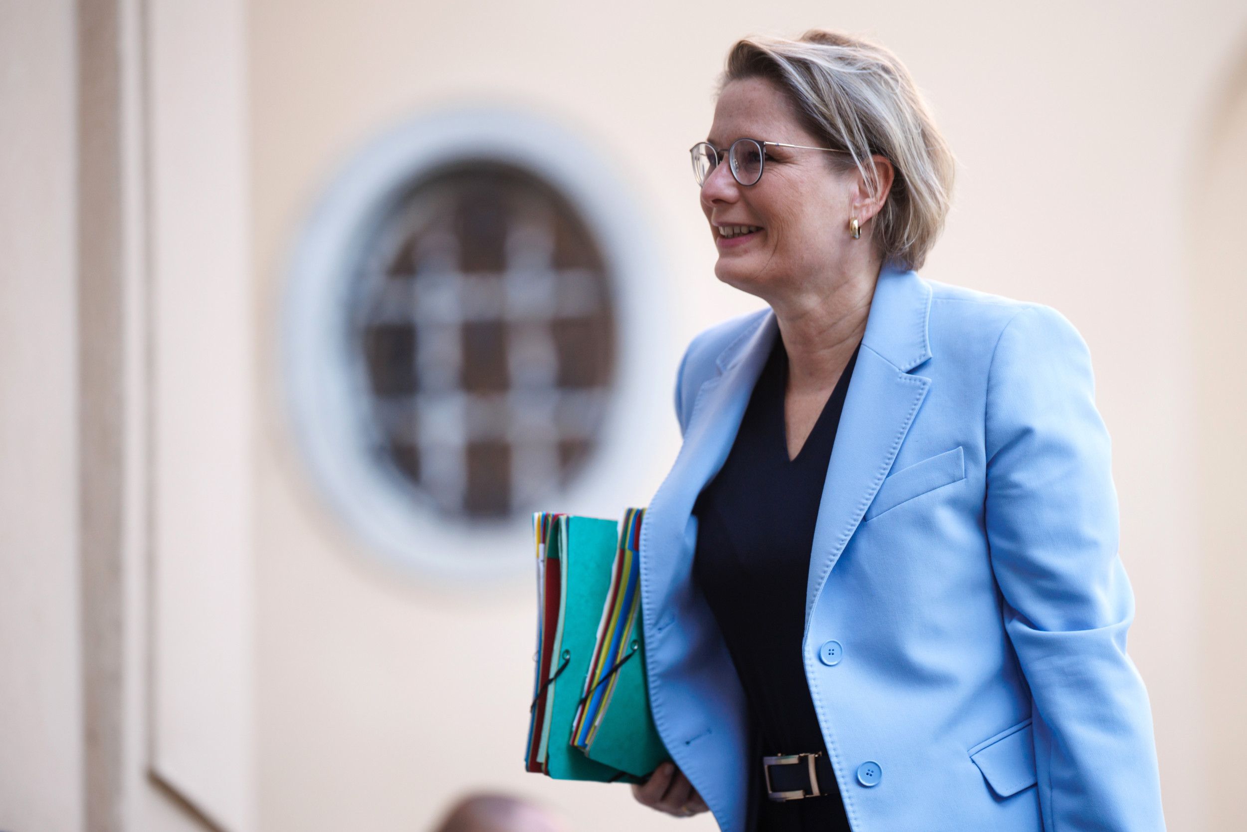 Een vrouw met bril en lichtblauw jasje loopt met mappen onder haar arm.