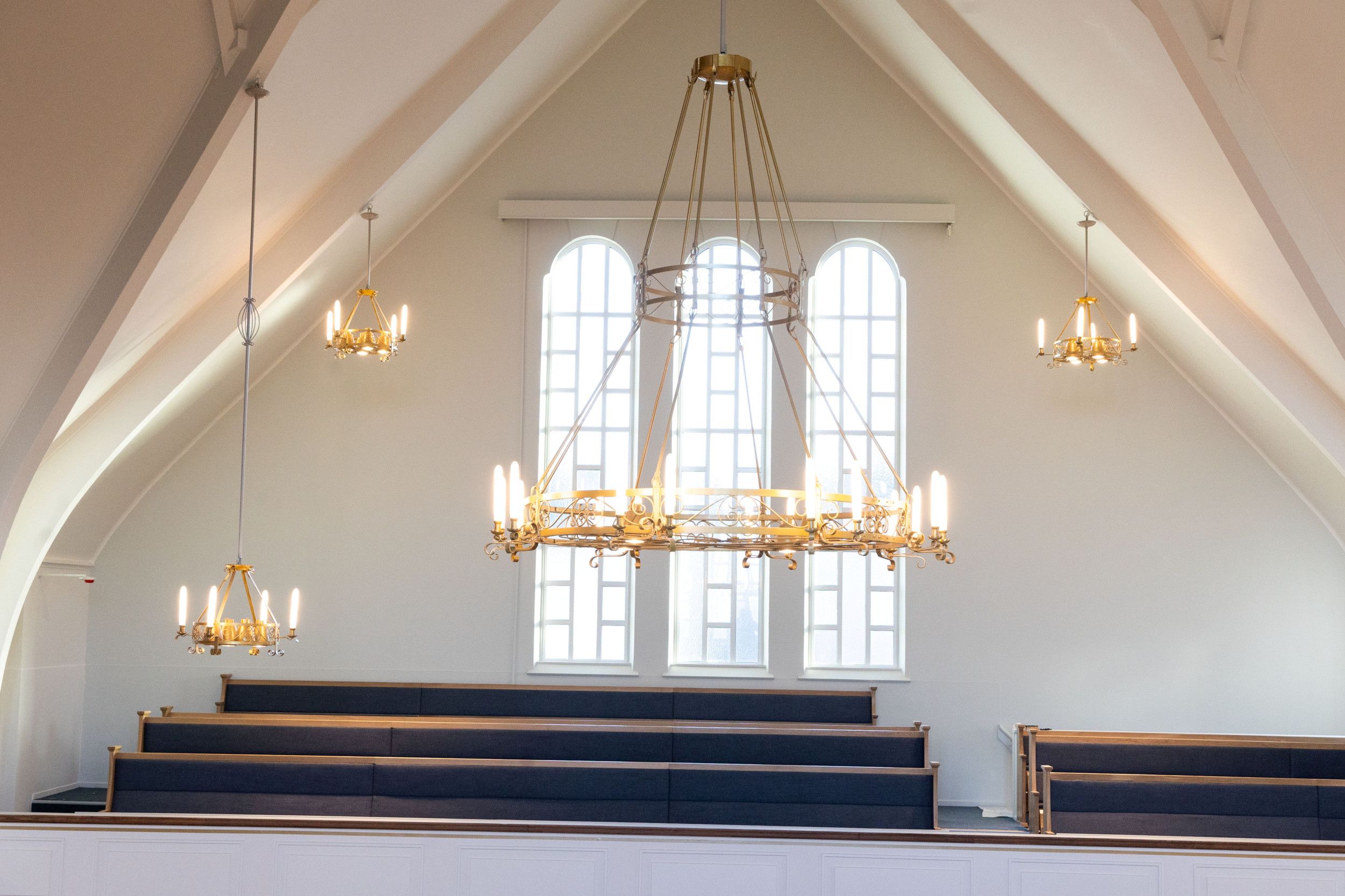 Wit interieur van een kerkgebouw met onderin donkere kerkbanken met erachter drie hoge boogramen en verschillende kroonluchters die vanaf het plafond naar beneden hangen.
