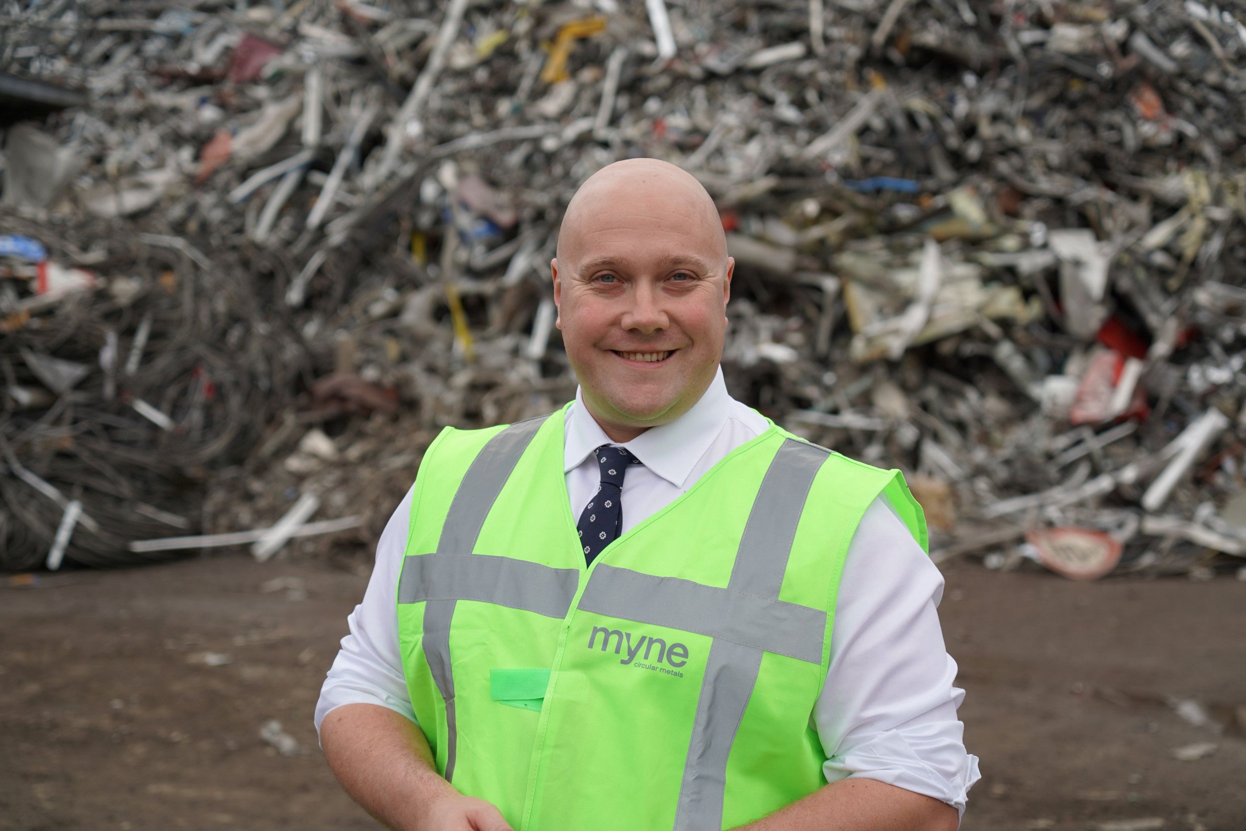 Glimlachende kale man met een geel hesje aan. Op de achtergrond een hoop schroot. 