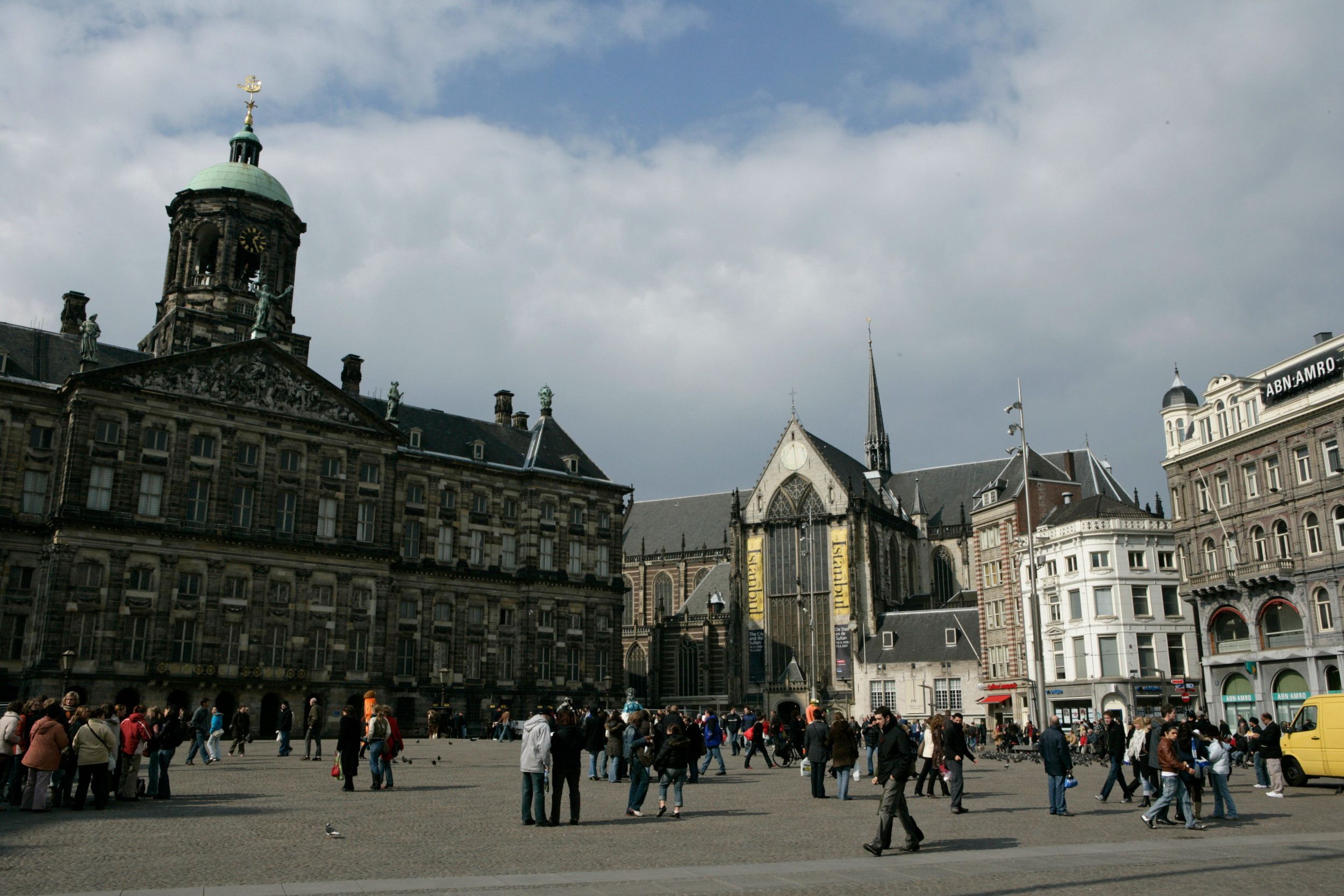 Foto van een stadsplein met links een monumentaal gebouw en rechts een kerk met een kleine spits op het dak. Er lopen veel mensen op het plein.