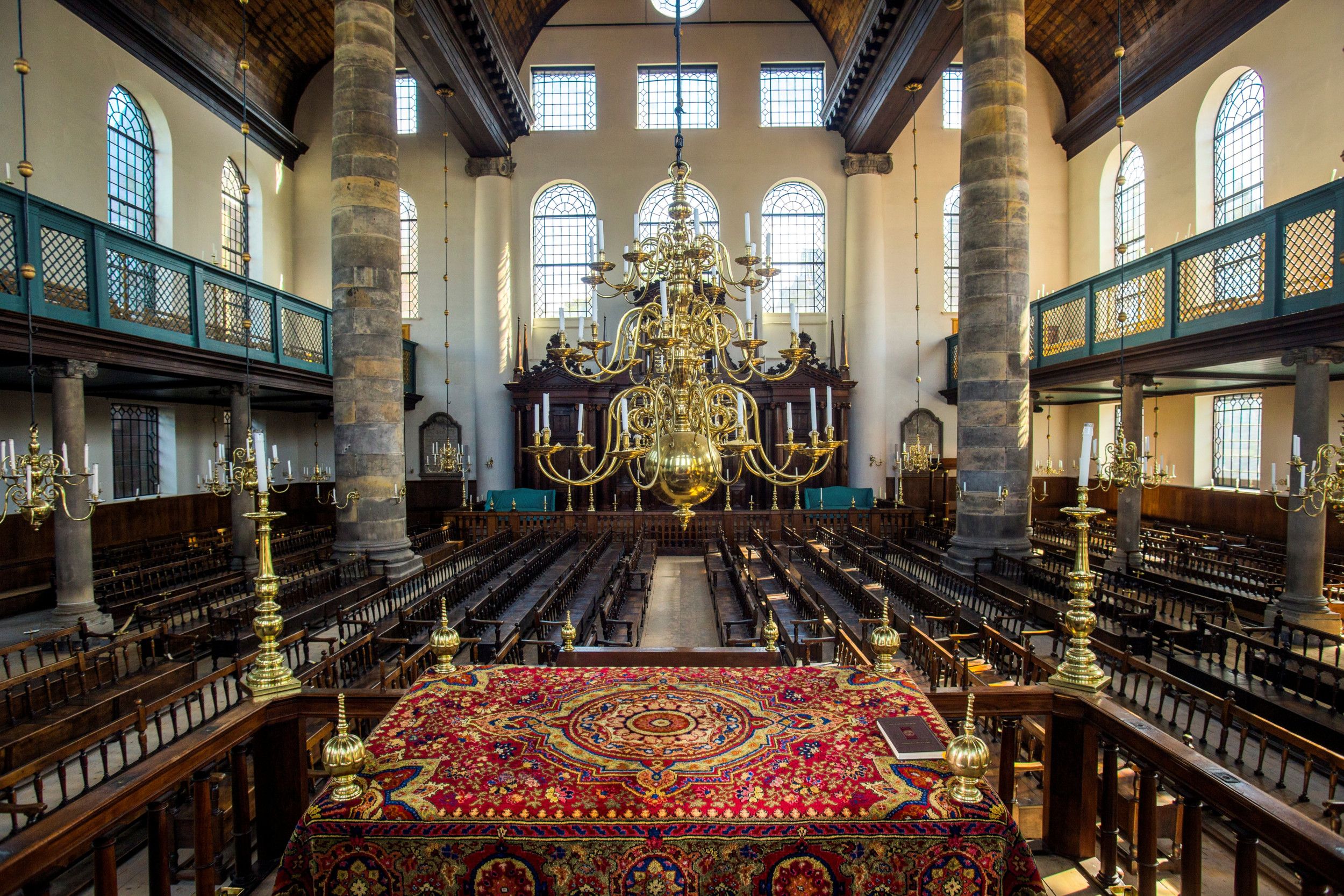 Foto van het interieur van een kerkelijk gebouw. In het midden rijen houten banken onder een kroonluchter. Links en rechts stenen pilaren en balustrades voor de ramen.