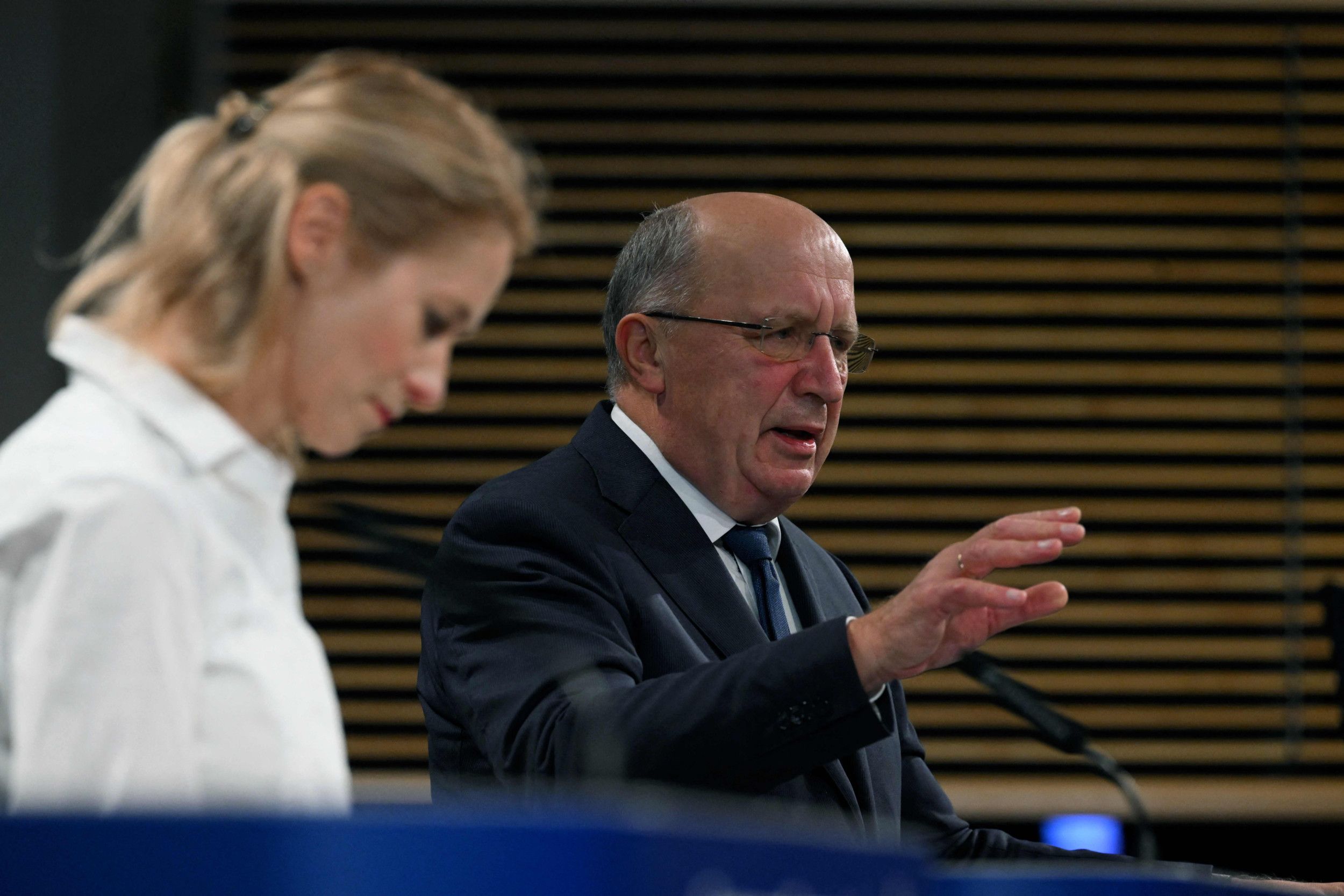 Een oudere man in pak met een bril op spreekt achter een spreekgestoelte. Links naast hem staat een jonge vrouw met blond haar in een witte blouse.