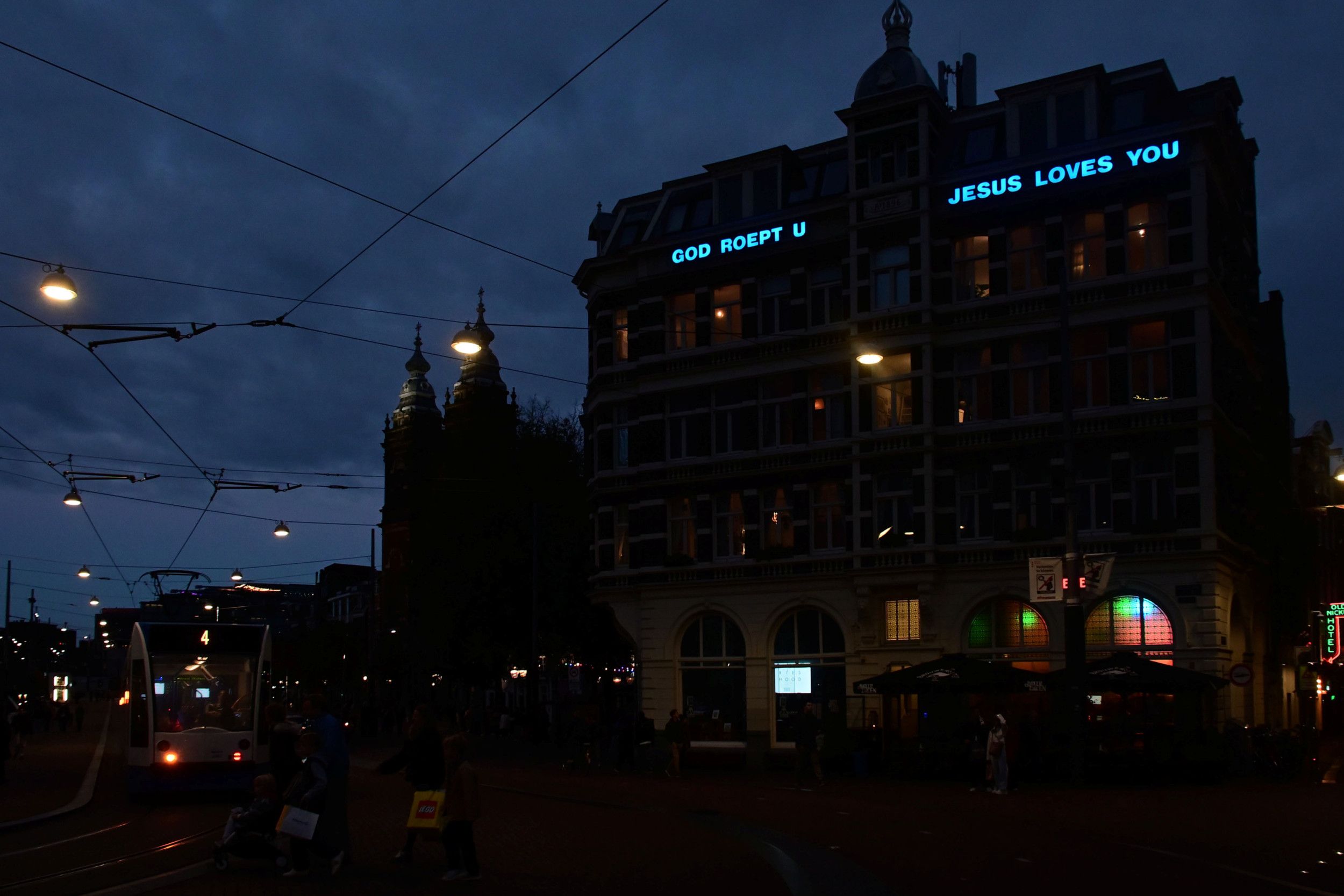 Stadsbeeld bij avondlicht. Tegen een donkerblauwe lucht zijn enkele monumentale panden te zien, onder meer een kerk met twee torens. Op het gebouw rechts staat in blauwe lichtgevende letters de tekst ”God roept u – Jesus loves you”.