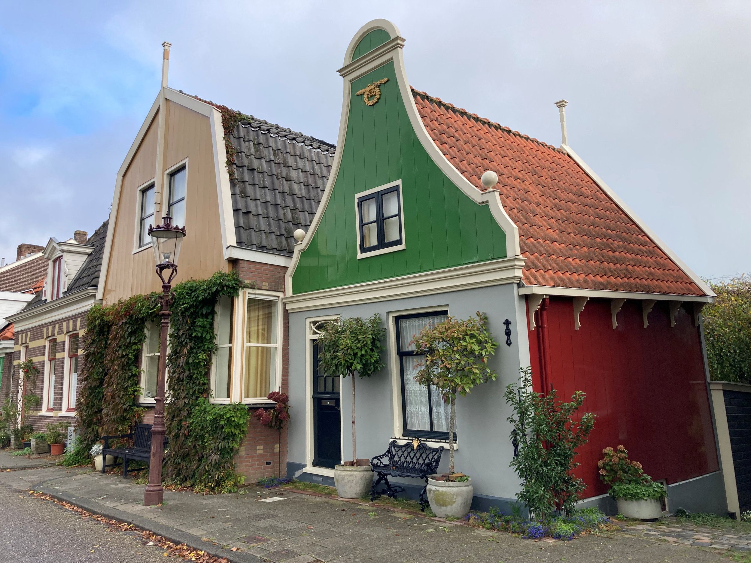 Twee Amsterdamse huizen, eentje met klokgevel en eentje met mansardekap. Het klokgevelhuis is bovenin groen. De voorgevel is grijs en de zijgevel rood.
