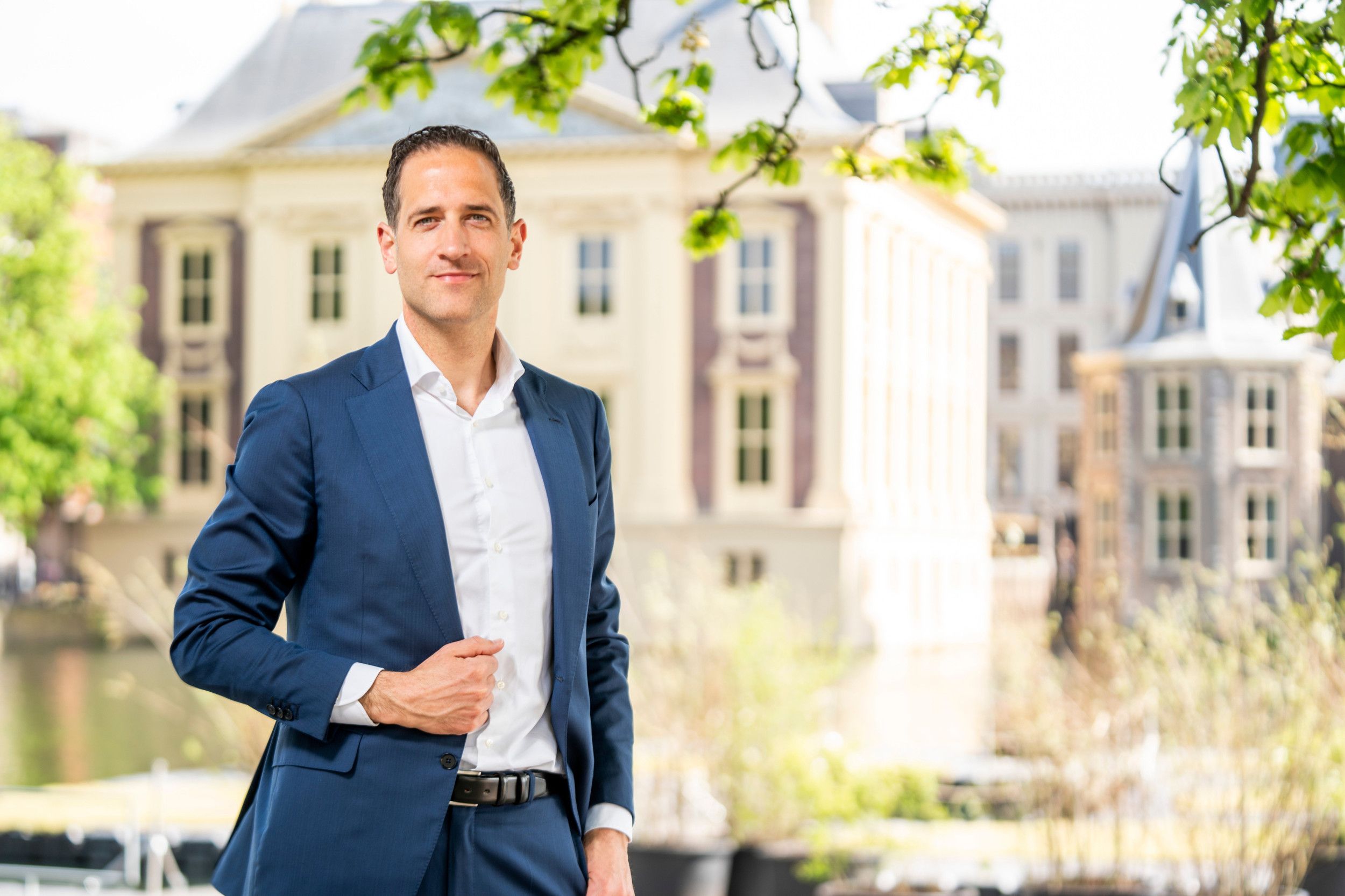 Jongeman met wit overhemd en blauw kostuum. Op de achtergrond gebouwen van het Binnenhof.