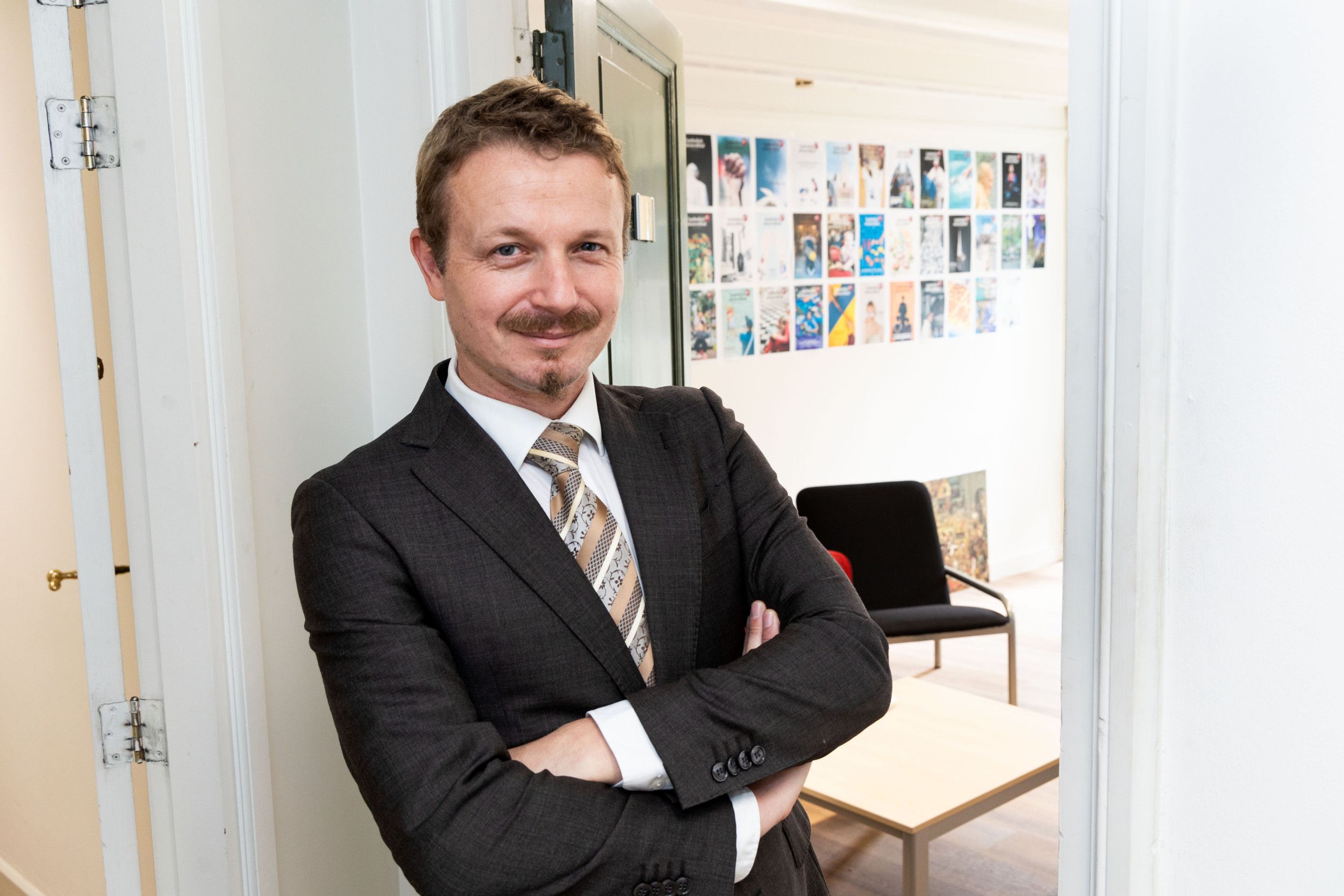 Man met bruin haar en snor in pak met stropdas met zijn armen over elkaar.