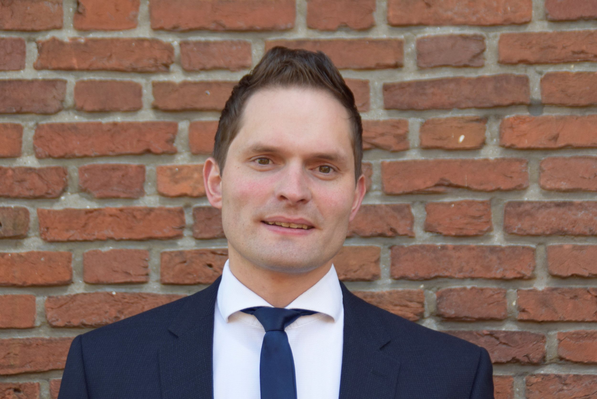 Man, glad geschoren, haar in een kuif gekamd, in donkerblauw pak met blauwe das, tegen de achtergrond van een bakstenen muur. 