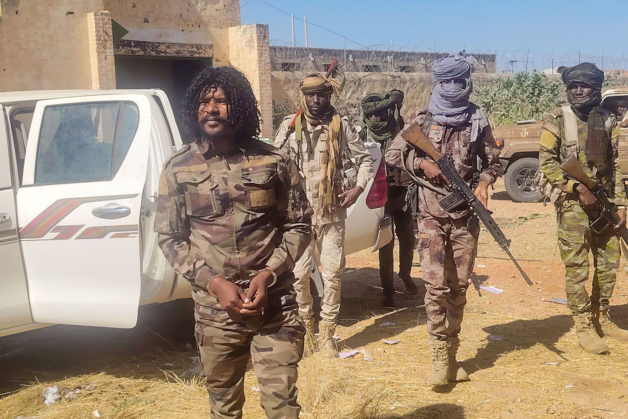 Mannen in groene camouflagekleding en met geweren staan achter een man met handboeien.