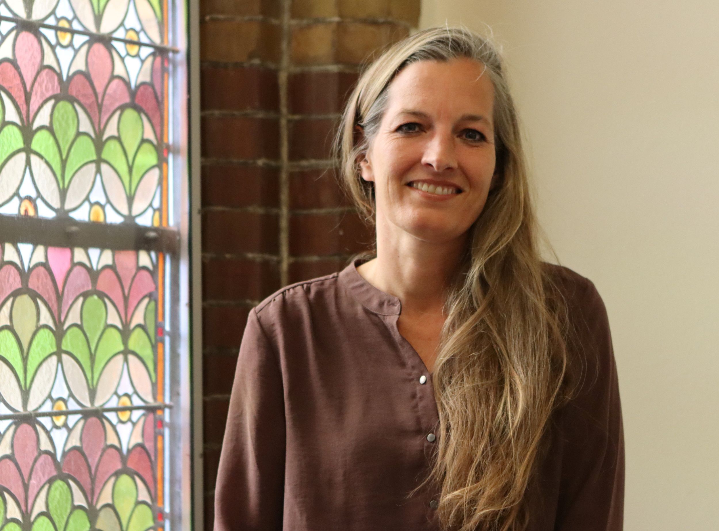 Vrouw met lang blond haar en bruine jurk naast een gebrandschilderd raam.