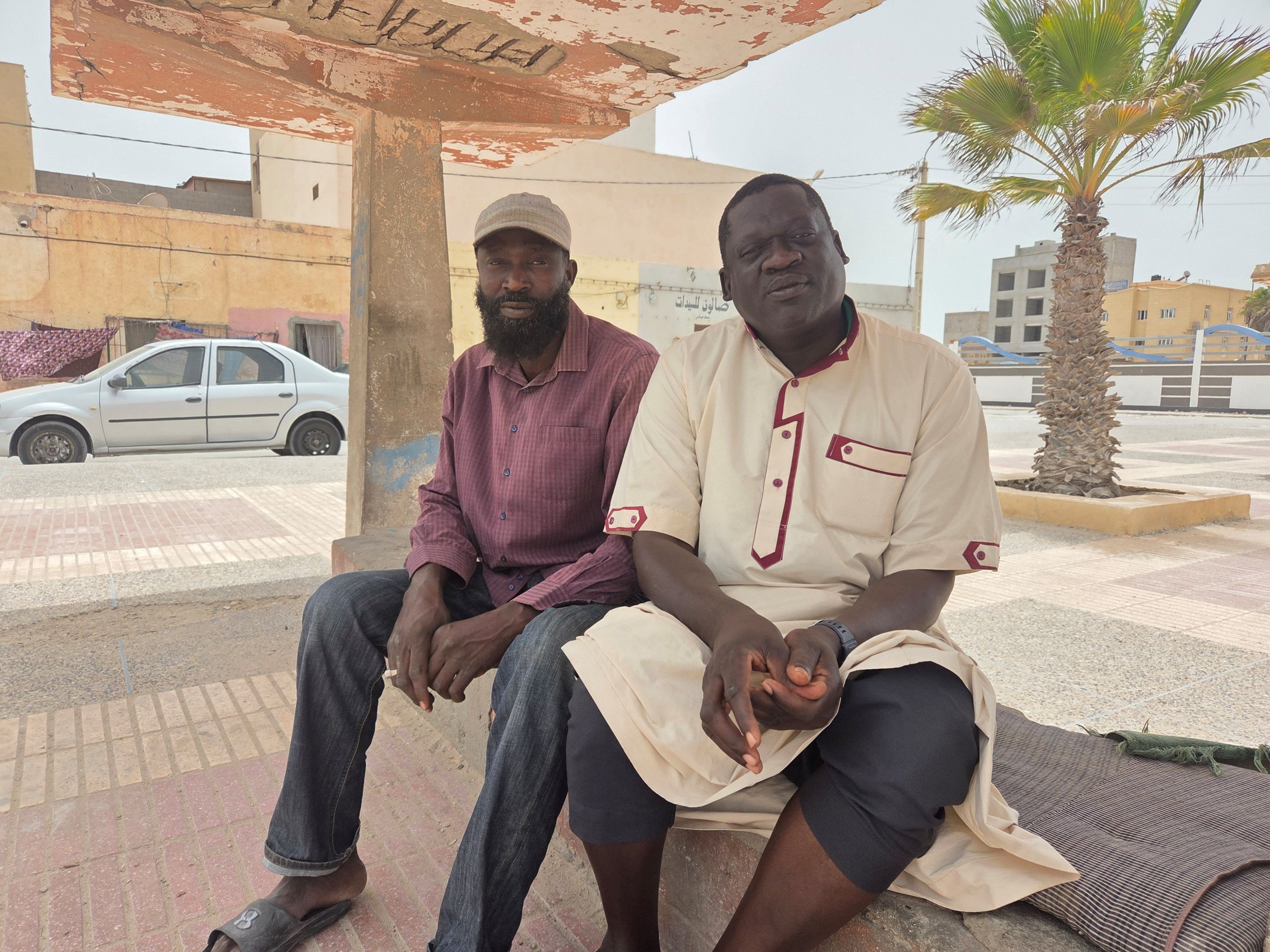 Twee mannen zitten op een betonnen bankje in de schaduw. 
