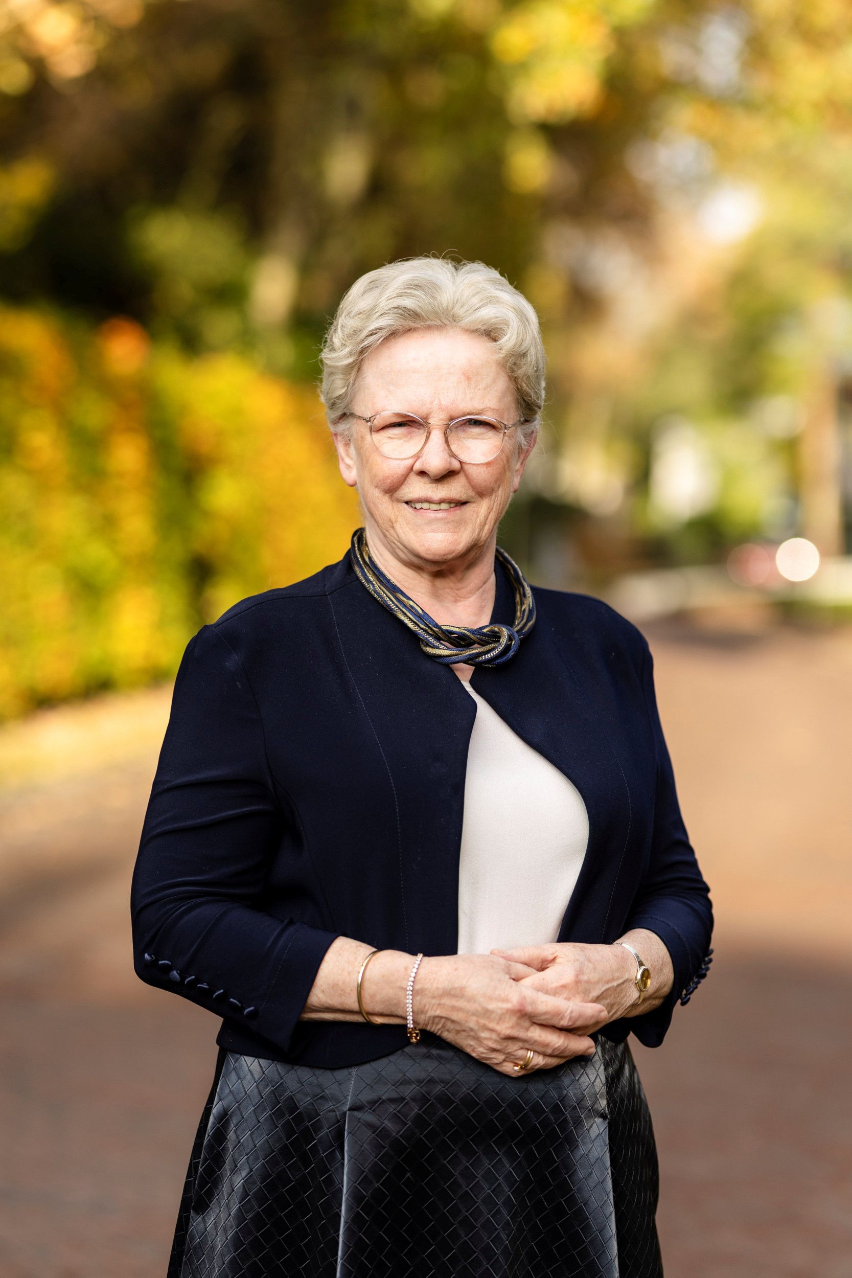 Oude mevrouw met grijs, opgestoken haar, montuurloze bril, glimlach, donker jasje, collier, de handen over elkaar.