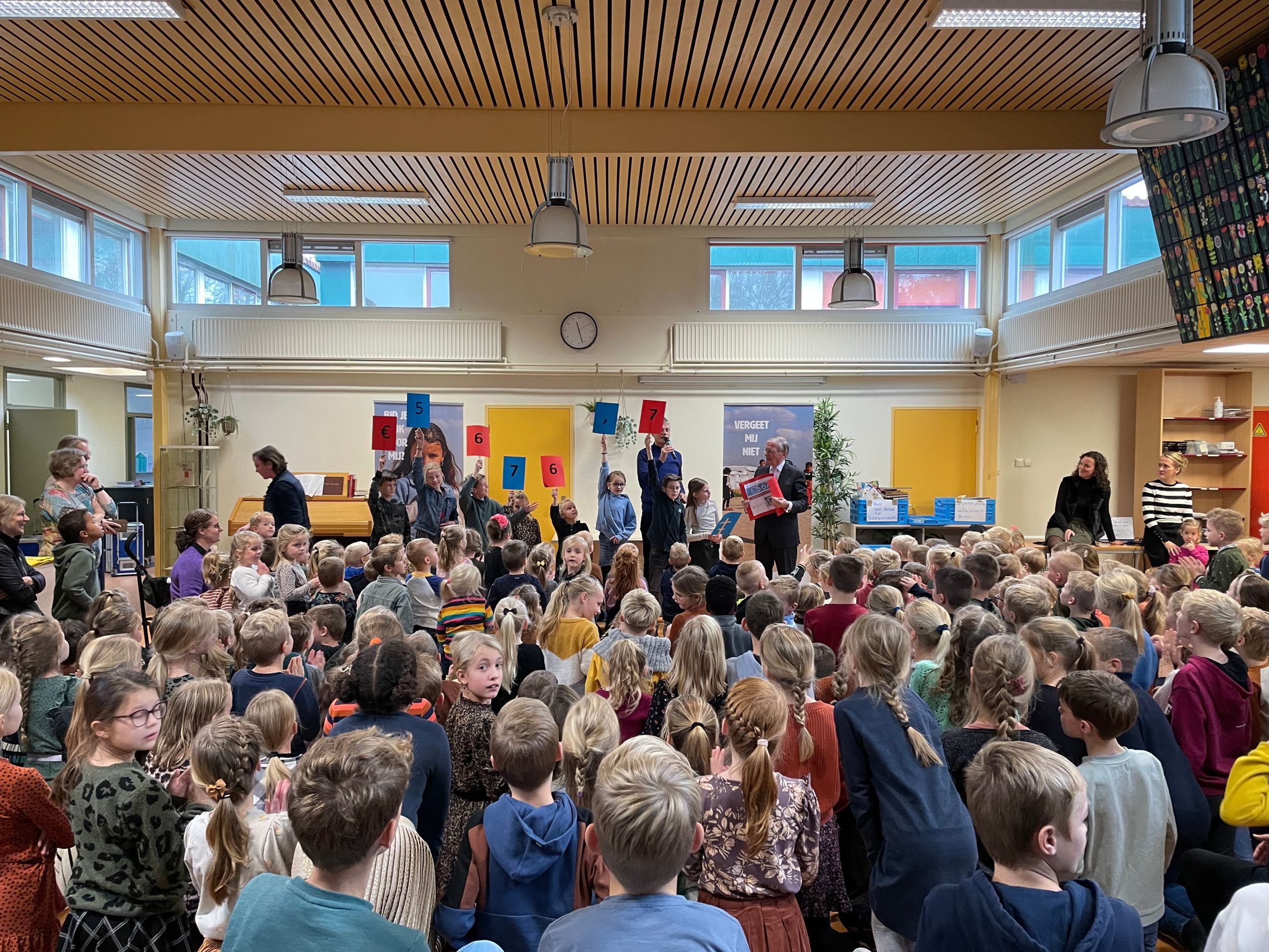 Basisschoolkinderen kijken naar een presentatie van de opbrengst van een actie. Kinderen op een podium houden bordjes met cijfers omhoog die het opgehaalde bedrag aanduiden: €5676. 