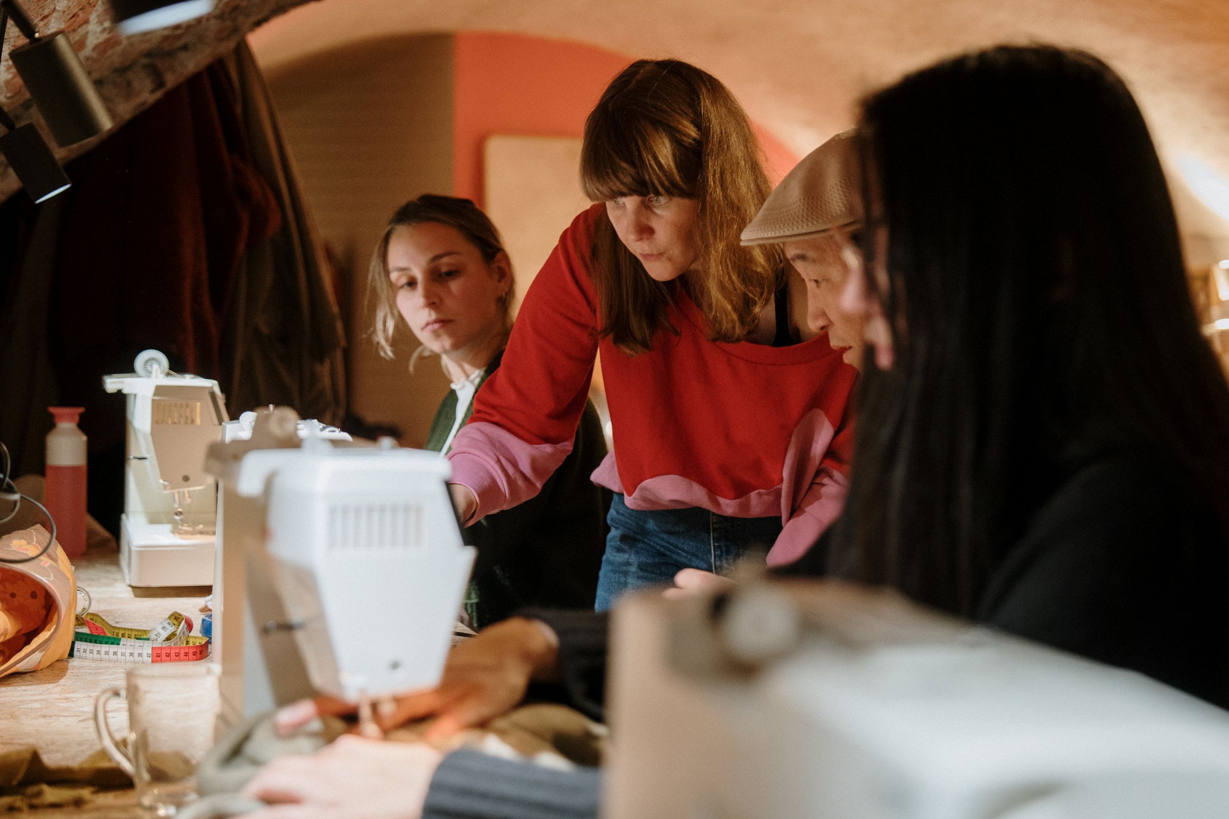 Een vrouw in rode trui wijst iets aan bij een naaimachine waar een Aziatische man met beige pet achter zit. Een vrouw met lang, bruin haar kijkt toe.  