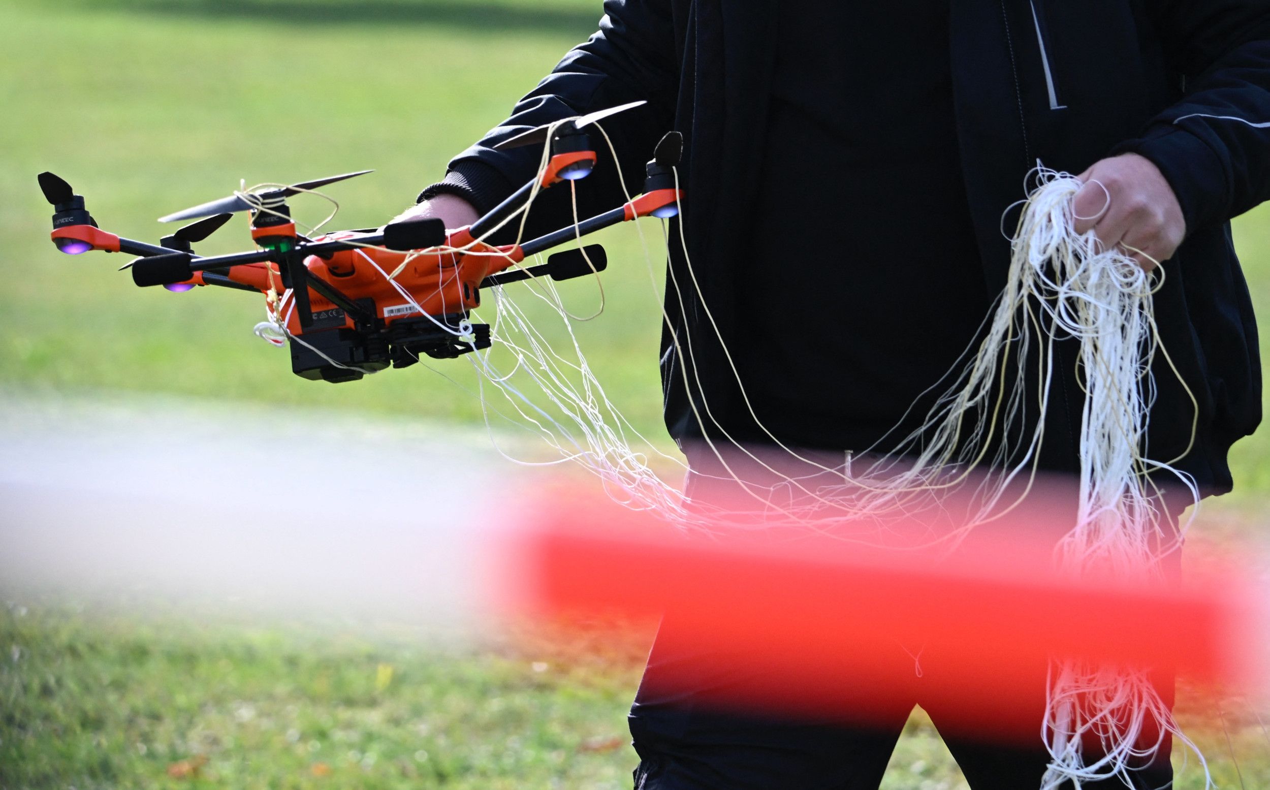 Een in het donker geklede persoon houdt in zijn ene hand een oranje drone vast, die in de klit zit met een wit net. De resten van dat net houdt de persoon in zijn andere hand.